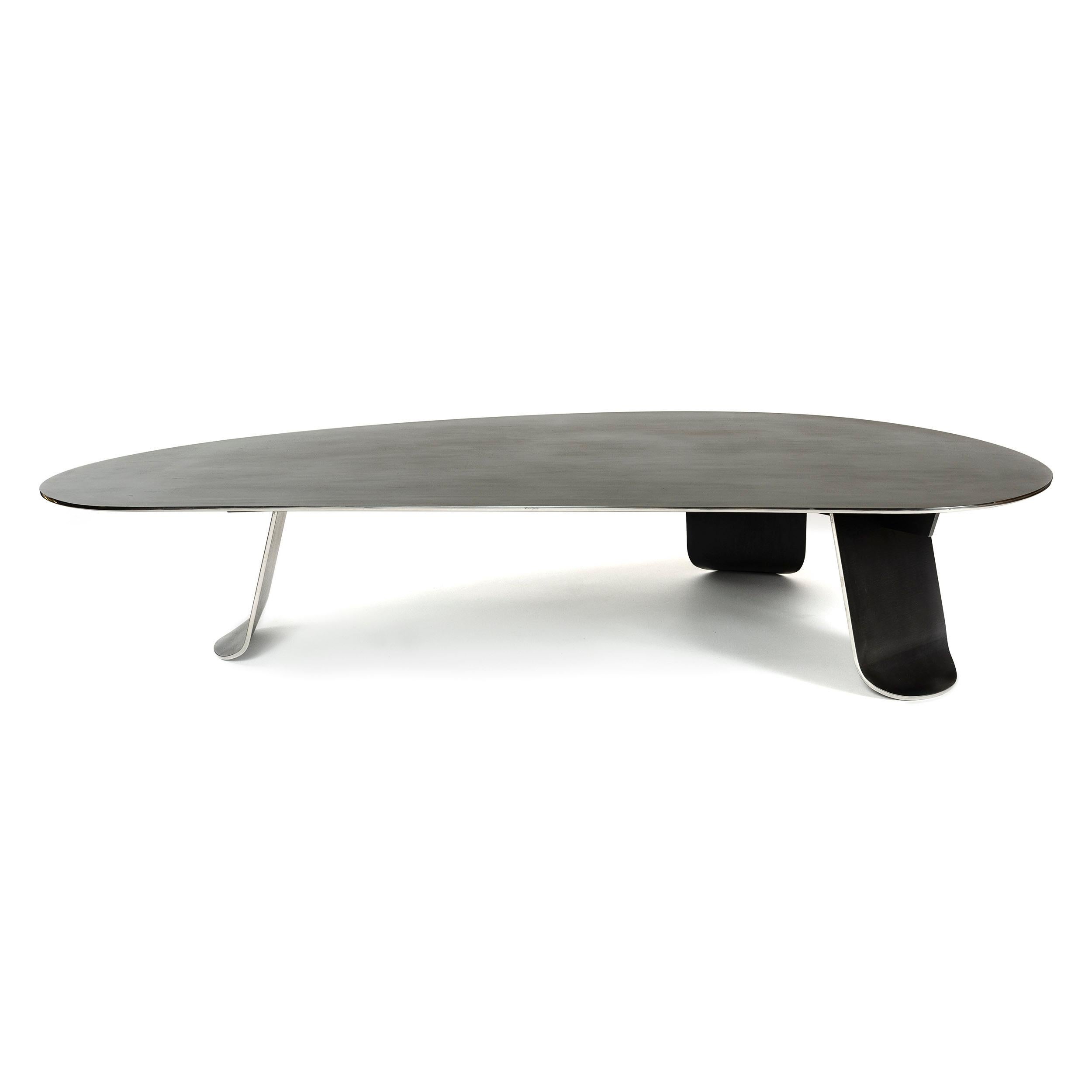A low coffee / cocktail table of thick, plate steel patinated a low sheen black with a polished edge. The expansive, organically shaped top is set upon three wide, thick plate steel legs each having gradually tapering sides terminating in softened