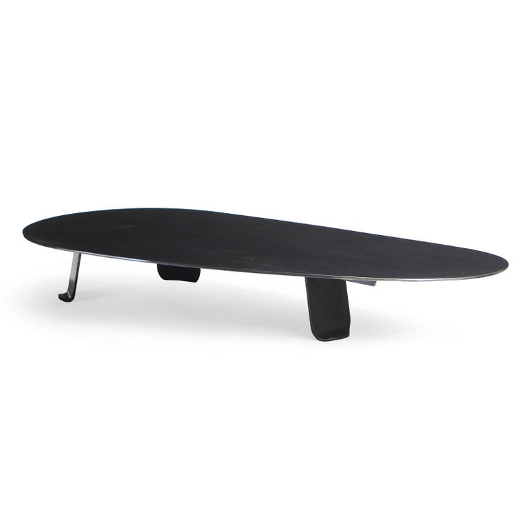 A low coffee / cocktail table of thick, plate steel treated with a blackened finish. The expansive, organically shaped top is set upon three wide, thick plate steel legs each having gradually tapering sides terminating in softened corners and