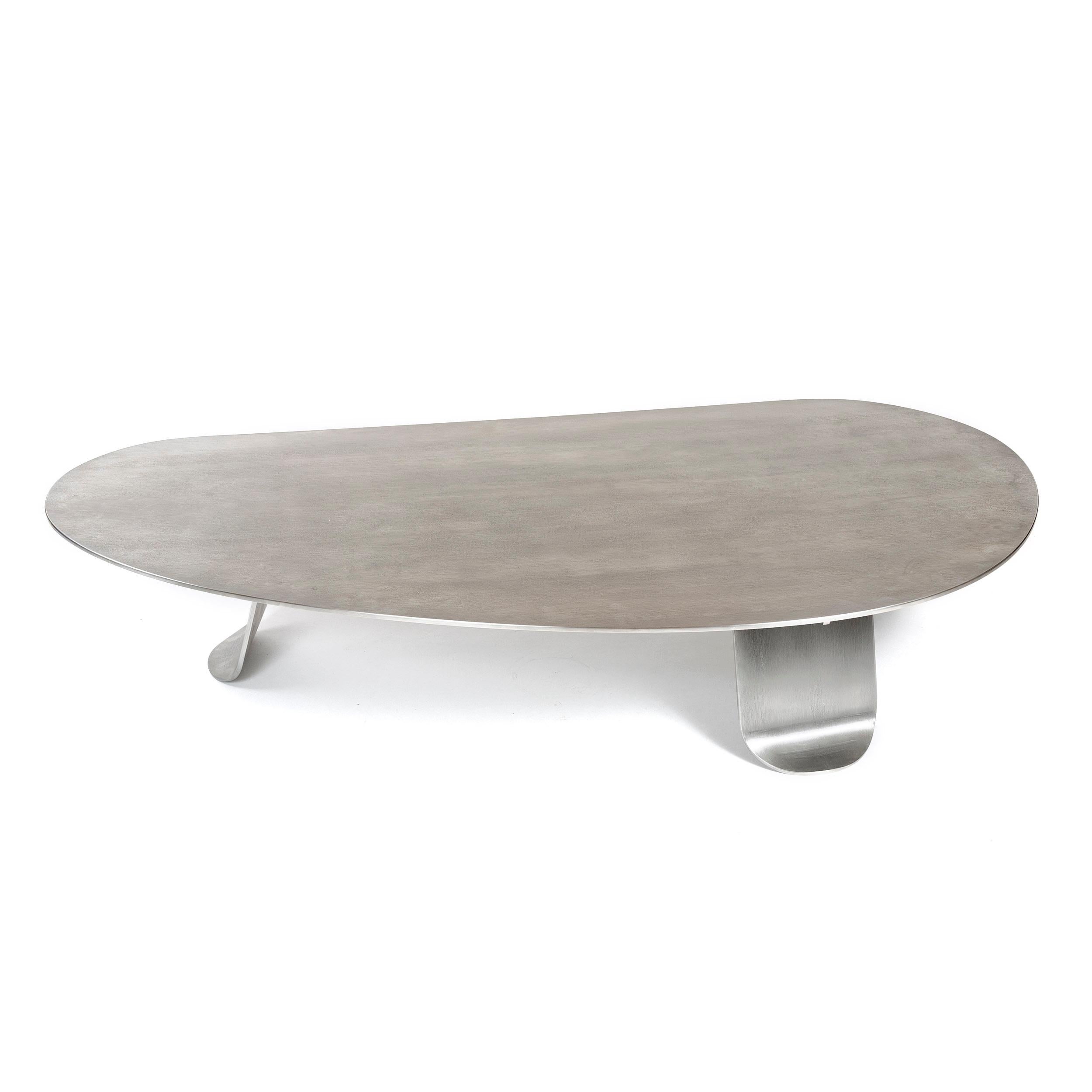 A low coffee/cocktail table of thick, plate natural grain stainless steel. The expansive, organically shaped top is set upon three wide, thick plate steel legs each having gradually tapering sides terminating in softened corners and subtle,