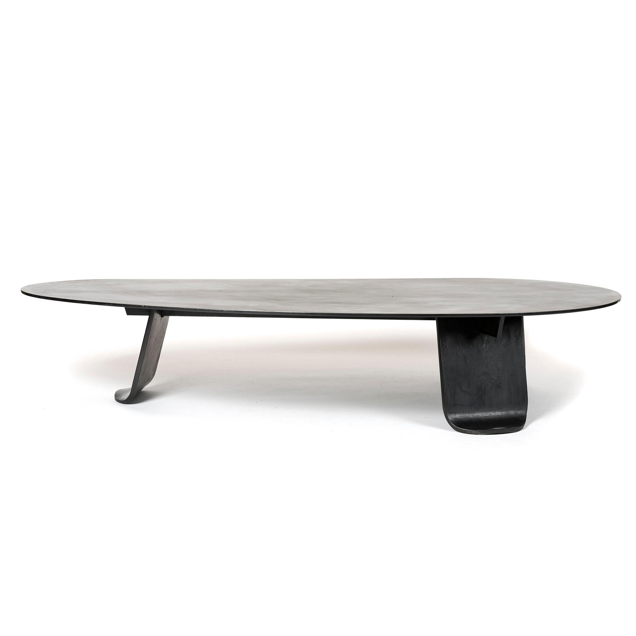 A low coffee / cocktail table of thick, plate steel treated with hot zinc and patinated a low sheen black. The expansive, organically shaped top is set upon three wide, thick plate steel legs each having gradually tapering sides terminating in