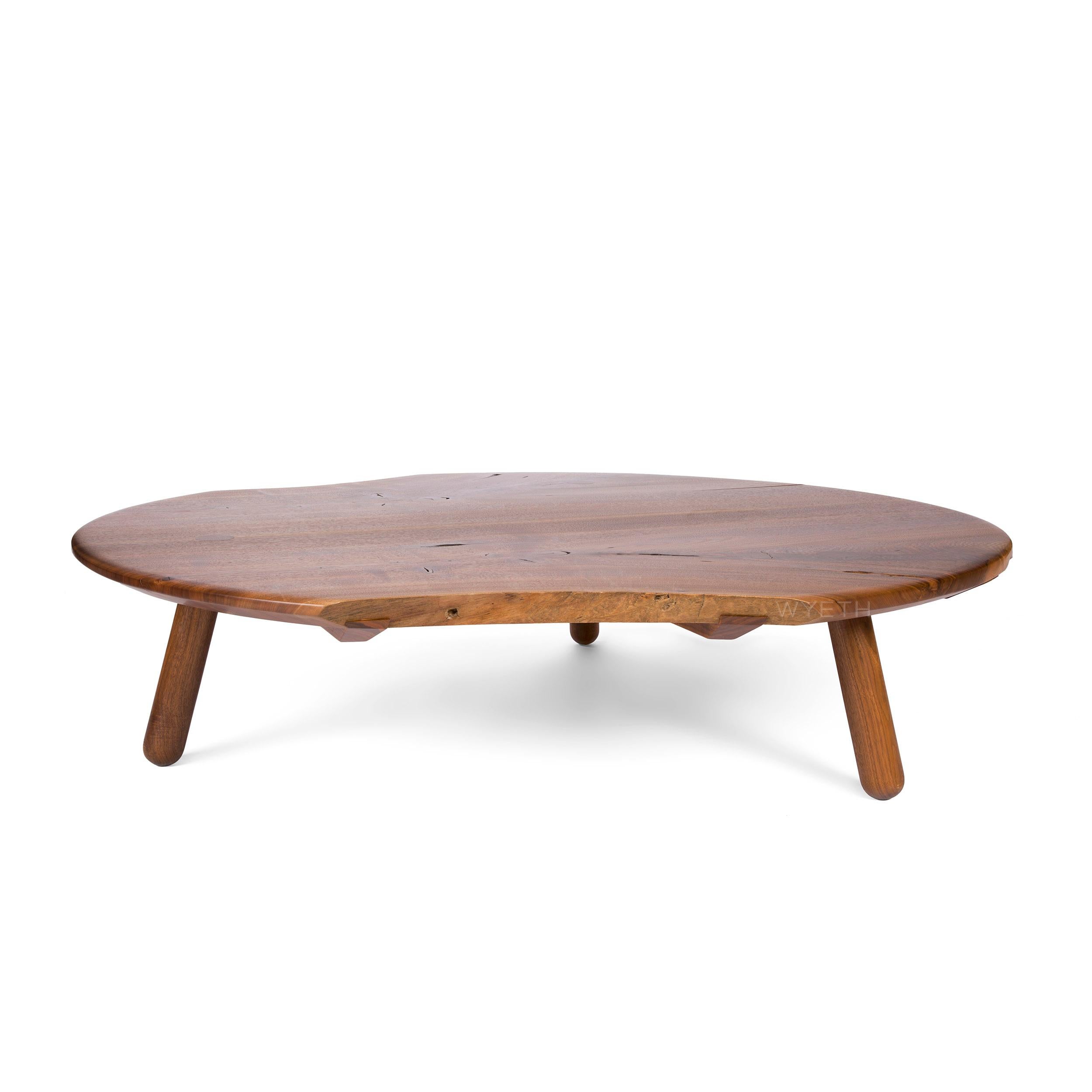 Ein handgefertigter, niedriger Tisch aus Nussbaumholz mit runder Form, natürlichen, dem Holz innewohnenden Kanten und drei (3) massiven, gedrechselten Beinen, die durch die Platte hindurchgeführt sind. Die Platte besteht aus lebenden Kanten und