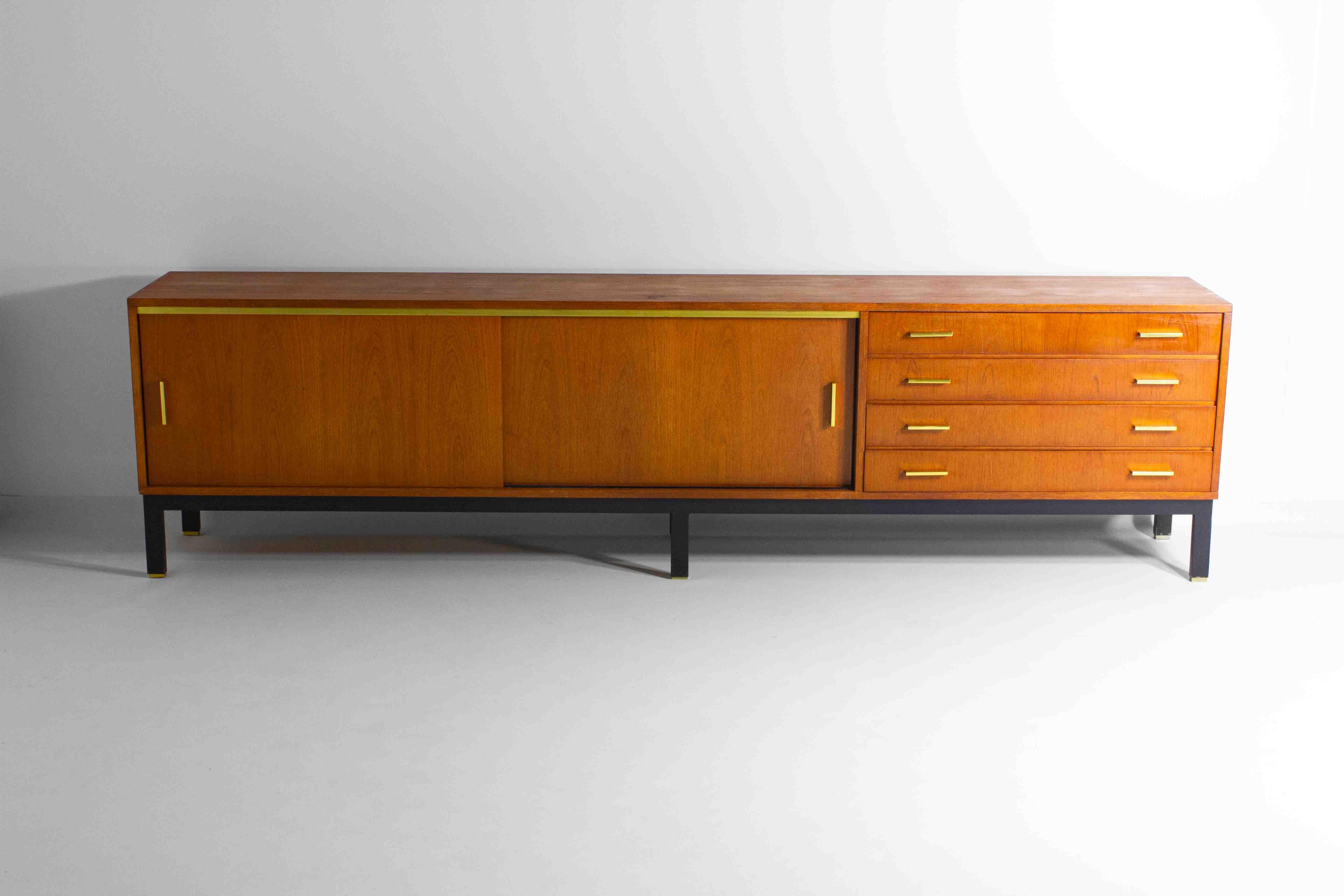 This sleek extra large sideboard is a typical mid-century beauty. Adorned in premium teak and finished with beautiful golden handles, sabots and sliding bars. The firm wooden corpus rests on a black metal base.