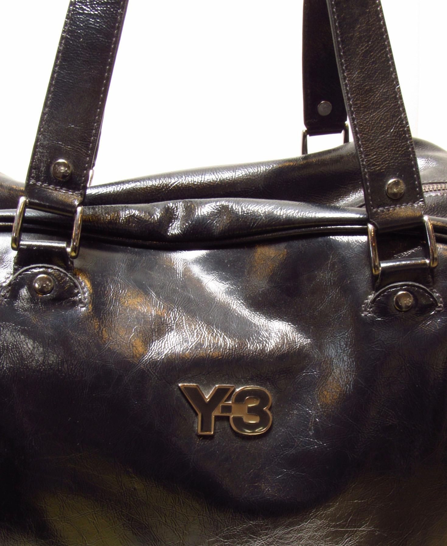 This sporty vintage black patent leather retro bowling bag features woven mocha brown panels, double zippers for ease of access, a discrete side pocket, and the Y-3 logo. The inside provides three slot pockets and one zippered pocket. 