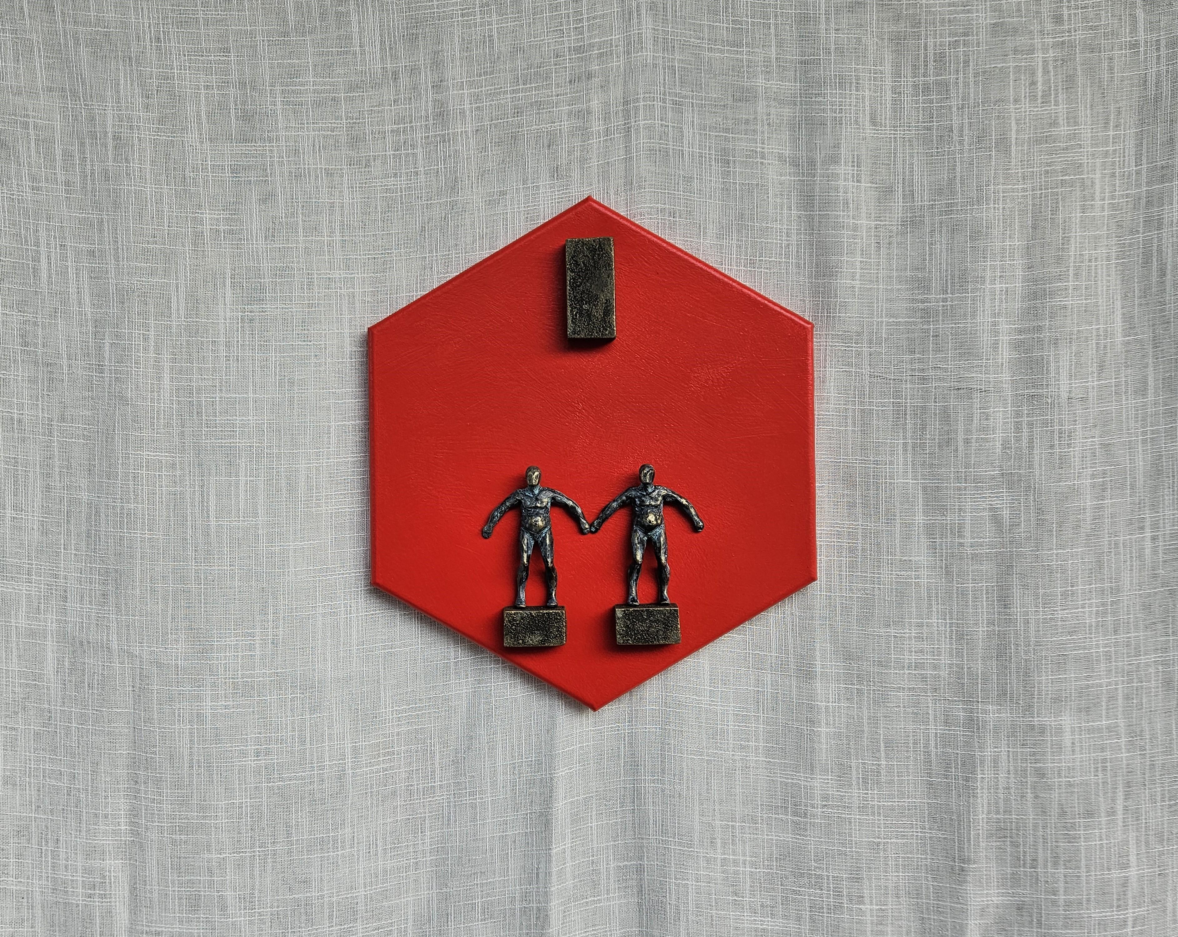 <p>Comentarios del artista<br>Dos figuras humanas están de pie, separadas, sobre una plataforma, con las manos tocándose. Un bloque rectangular flota sobre ellos. La base hexagonal roja refuerza la armonía visual, ya que infunde energía y calidez de