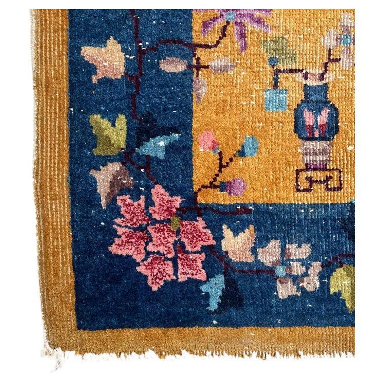 A colorful art deco chinoiserie rug in yellow and blue. This petite carpet has a yellow-orange background is surrounded by a deep blue border. It is decorated with pink, purple, blue, and green flowers throughout and accented with ceramic motif