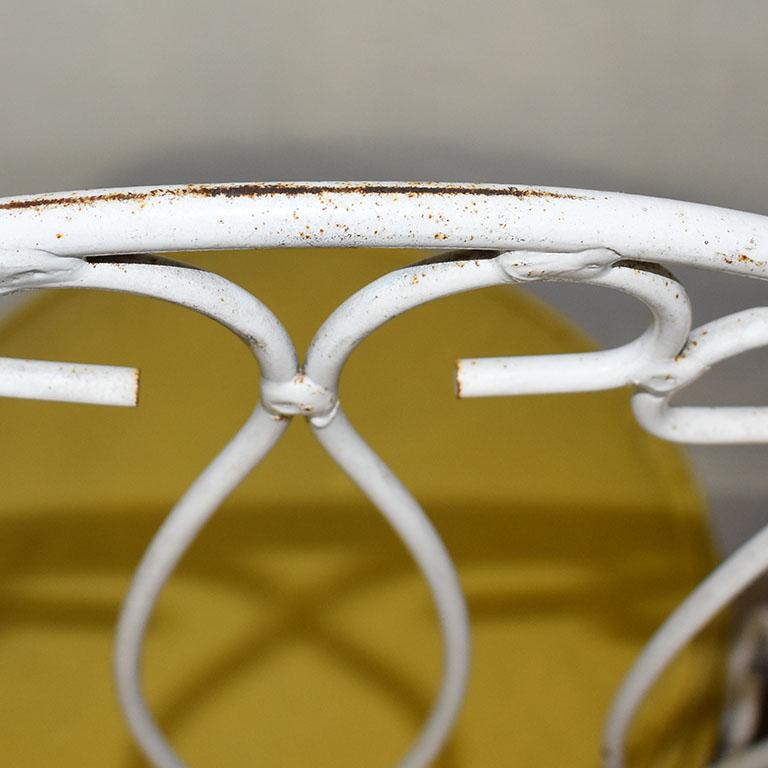 A pair of white iron patio chairs with round yellow vinyl seat cushions. This pair will be perfect in a garden setting. Each chair features a rounded back with stylized curled designs. The cushions are removable and upholstered in bright yellow