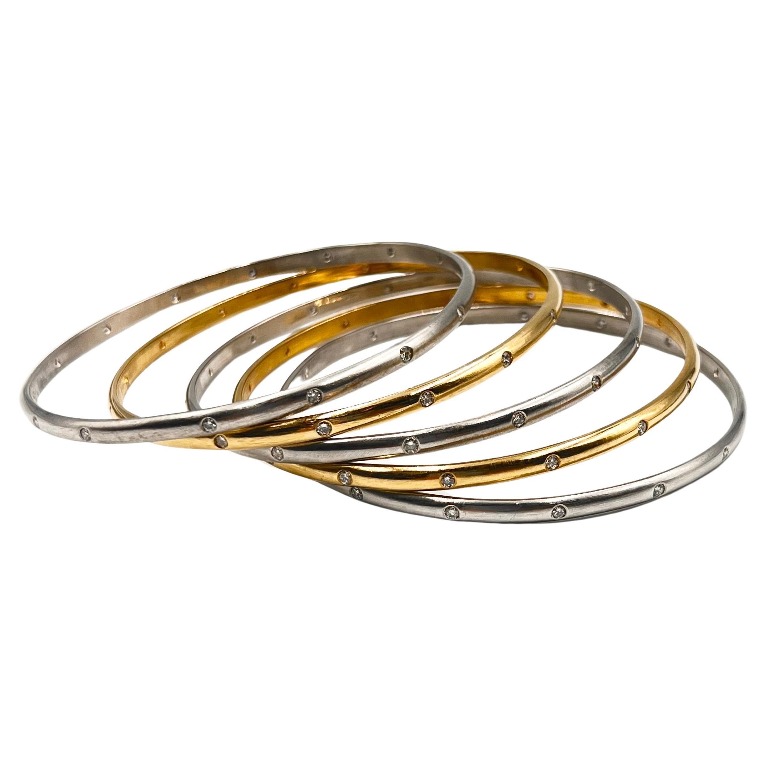 Five bangle bracelet set in polished 18k yellow gold and 14k white gold accented by round brilliant-cut diamonds. Two bracelets are in 18k yellow gold and three are in 14k white gold.  Each bracelet is bezel-set with fifteen round brilliant-cut