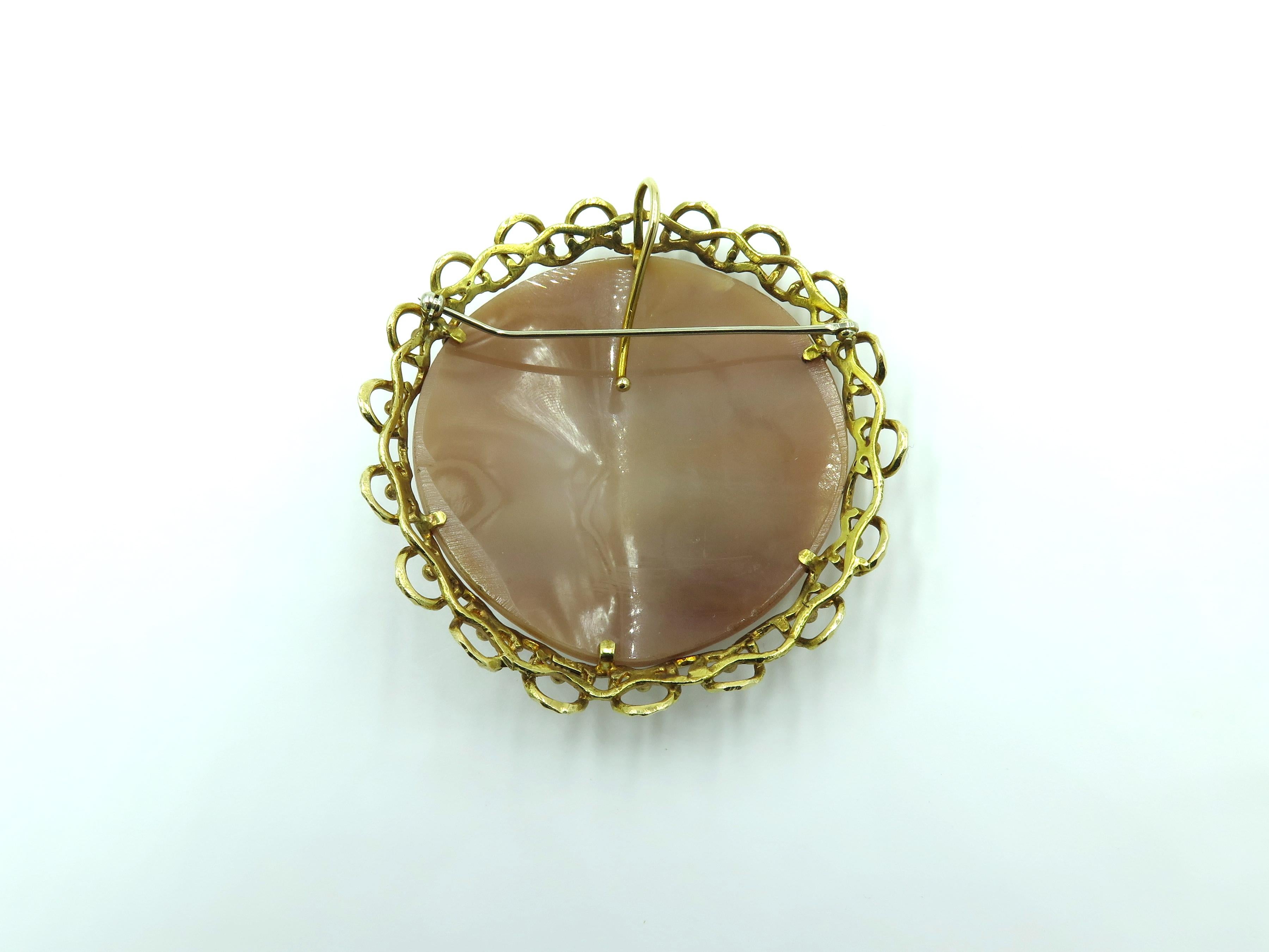 A 14 karat yellow gold and shell cameo brooch. Circa 1960. Centering a circular carved shell cameo depicting a scene from Classical Mythology, carved with woman holding a lyre with child, within a textured and openwork surround. With hoop for