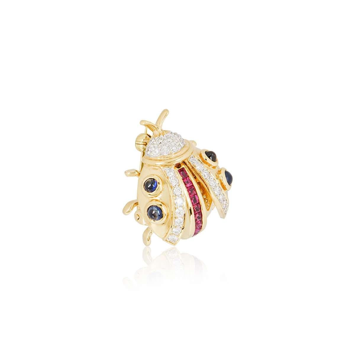 An 18k yellow gold Ladybird brooch. The brooch is set with 4 cabochon sapphires throughout the wings, with a row of 9 square cut rubies running through the centre of the body and 28 round brilliant cut diamonds set in the wings and head. The rubies