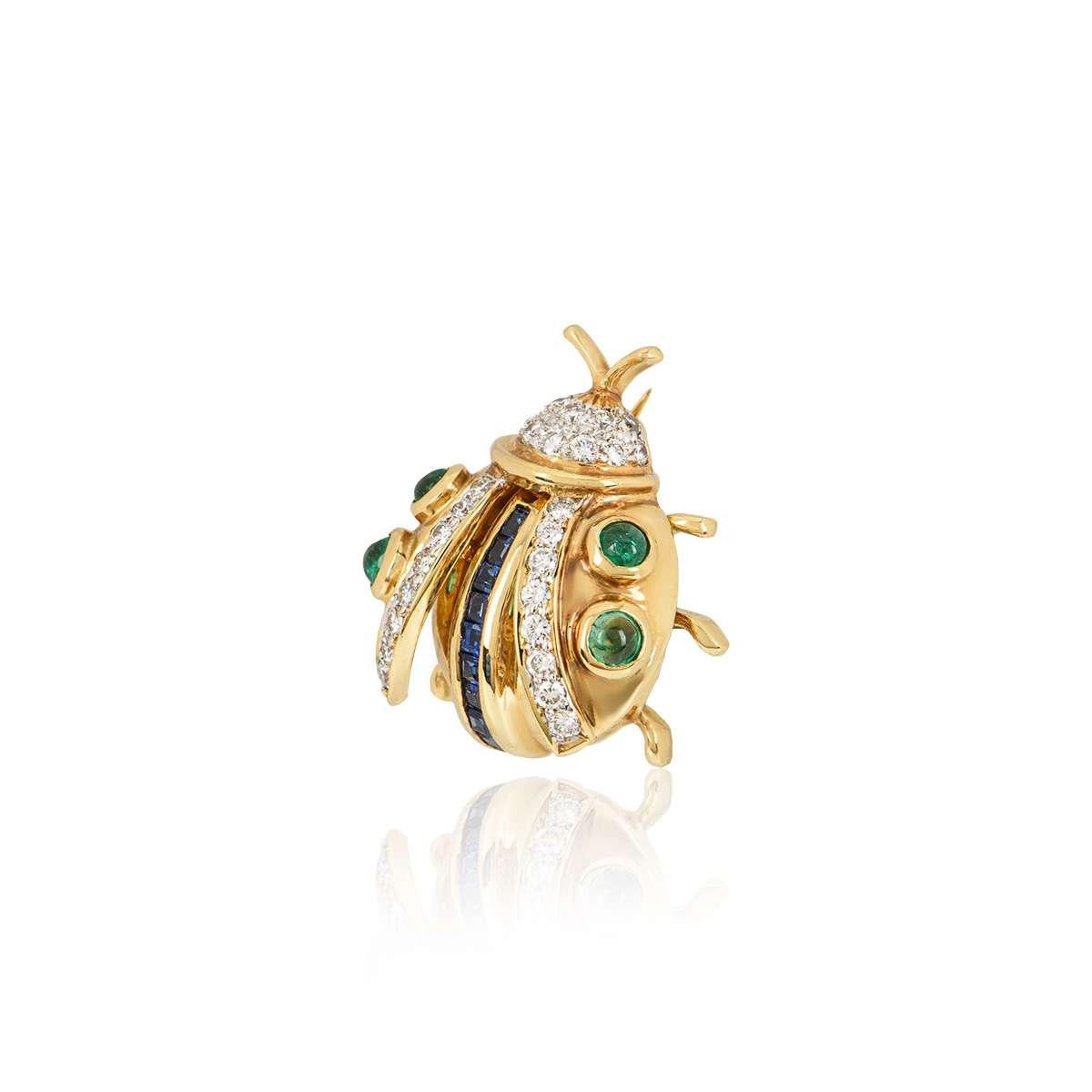An 18k yellow gold Ladybird brooch. The brooch is set with 4 cabochon emeralds throughout the wings, with a row of 9 square cut sapphires running through the centre of the body and 28 round brilliant cut diamonds set in the wings and head. The