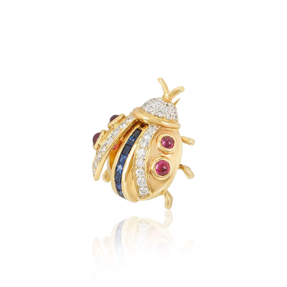 An 18k yellow gold Ladybird brooch. The brooch is set with 4 cabochon rubies throughout the wings, with a row of 9 square cut sapphires running through the centre of the body and 28 round brilliant cut diamonds set in the wings and head. The rubies