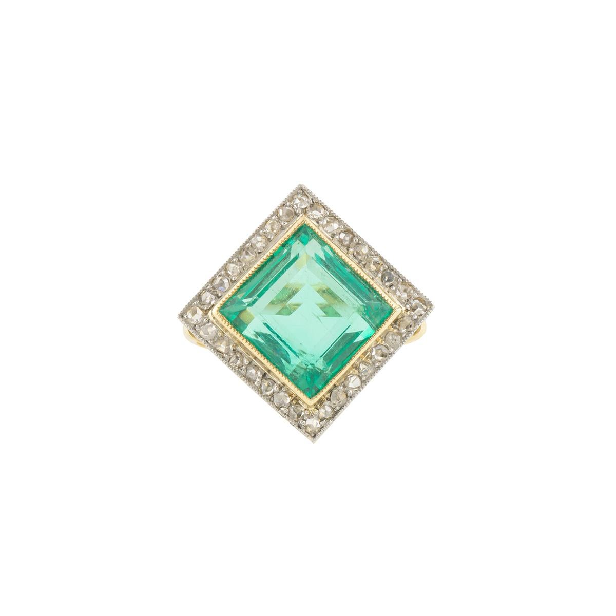 A luxurious 18k yellow gold emerald and diamond dress ring. The ring comprises of a princess cut emerald in a rubover setting approximately 5.50ct with a light green hue throughout. Complementing this are 32 rose cut diamonds with an approximate
