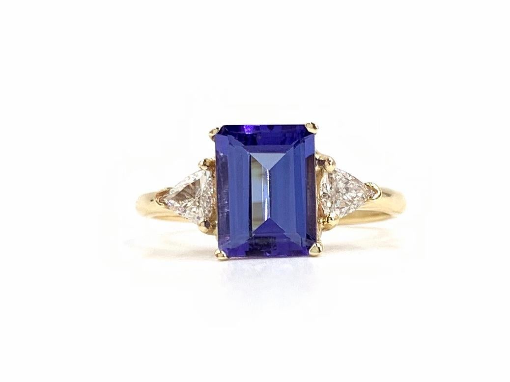 14 Karat yellow gold classic three stone style ring featuring a well-saturated violet hue Tanzanite with very nice clarity flanked by two trillion cut diamonds. Center emerald cut Tanzanite measures 8.5mm x 6.2mm at approximately 1.57 carats. White