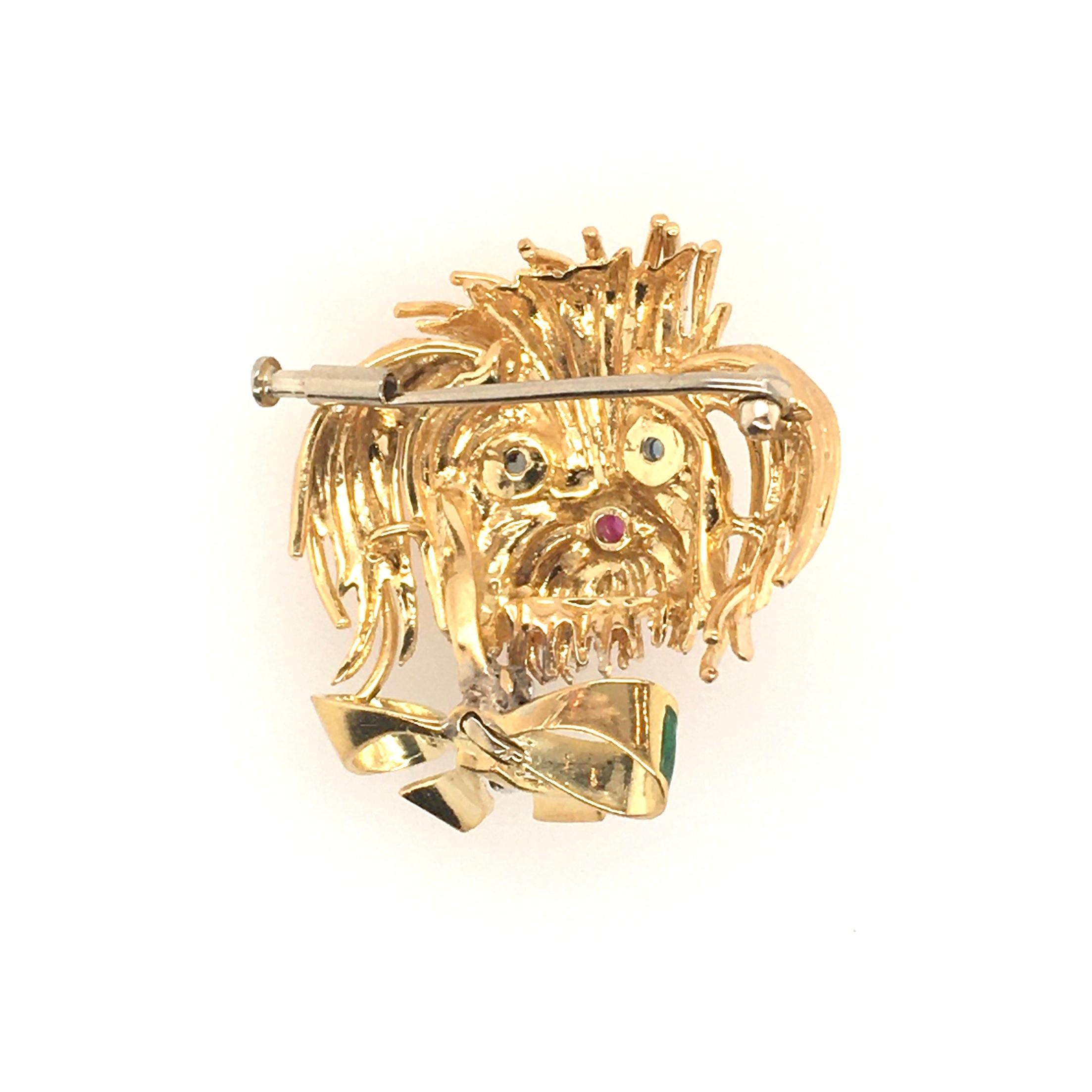 An 18 karat yellow gold, gemstone and enamel brooch. Designed as the head of a stylized Shih Tzu, with cabochon sapphire eyes and ruby nose, enhanced by a green enamel bow. Length is approximately 1 1/4 inches. Gross weight is approximately 11.3