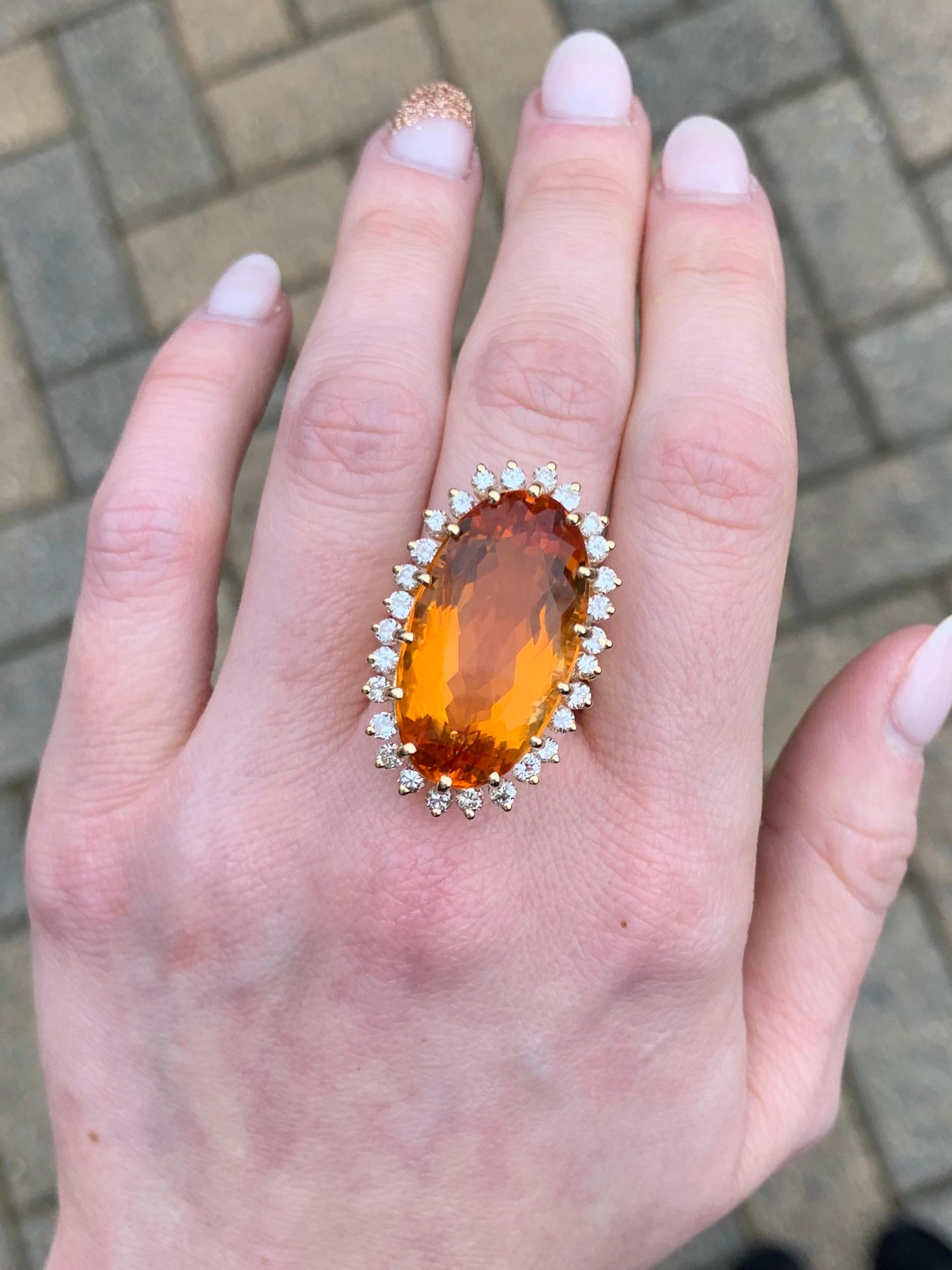 A timeless statement cocktail ring featuring a beautifully faceted large oval citrine topaz surrounded by a halo of prong set round brilliant diamonds. Citrine measures 24mm x 13.50mm. 28 round brilliant diamonds have an approximate total weight of