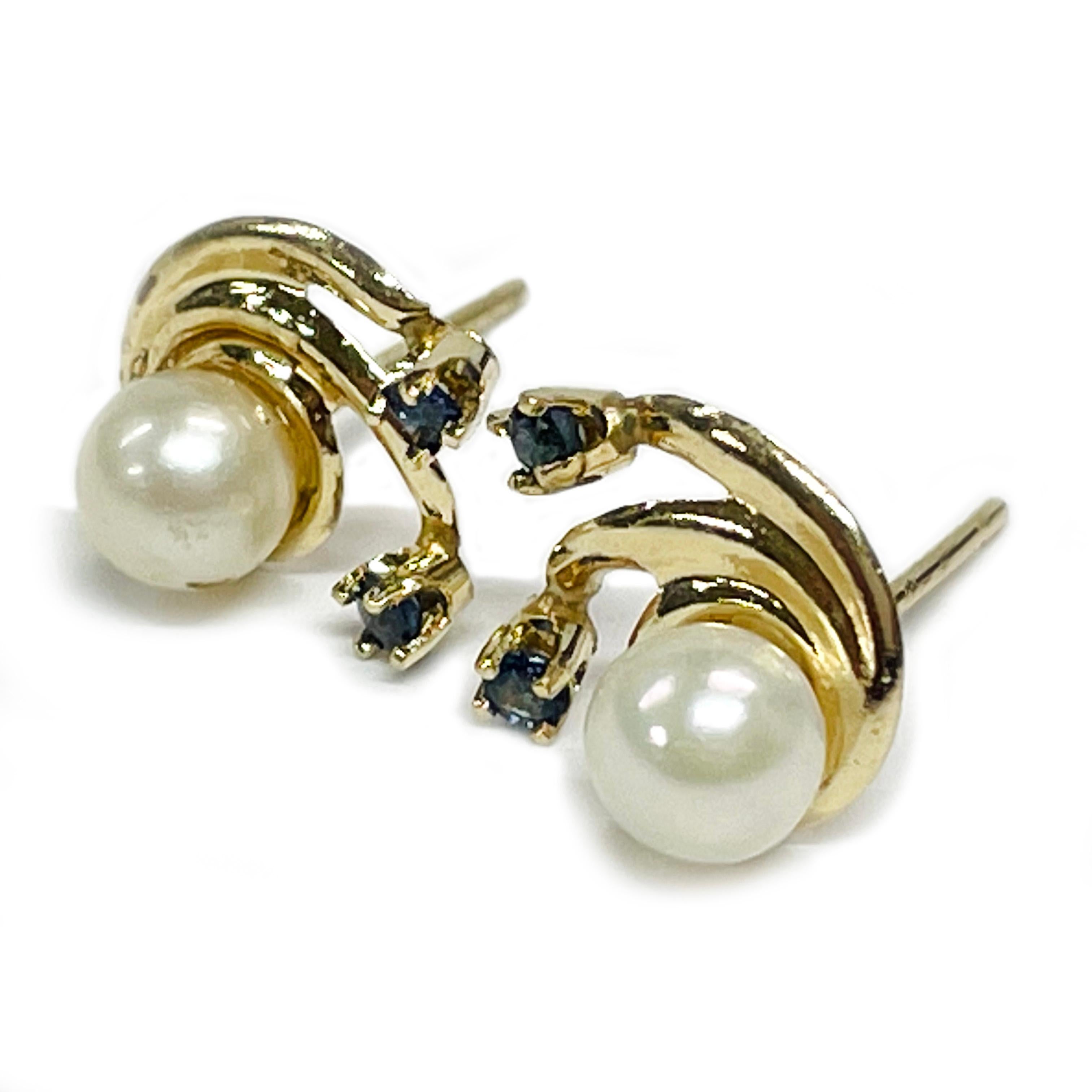 14 Karat Yellow Gold Pearl Blue Sapphire Stud Earrings. Each earring features a 4.75mm white pearl with two blue sapphires prong-set at the end of gold semi-circle swooshes. The pearls have a cream colored tone and both have good luster. The round