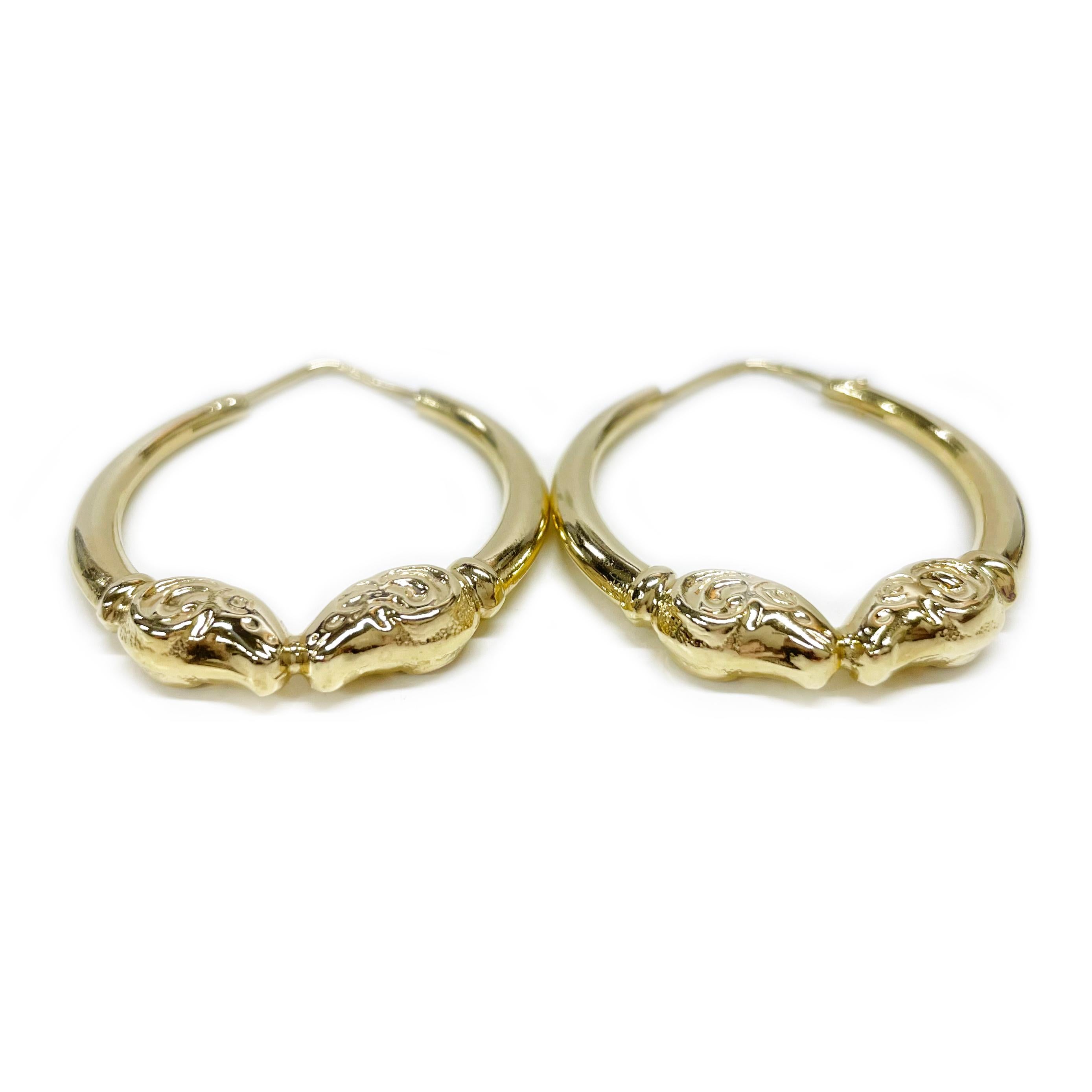 14 Karat Yellow Gold Ram Hoop Earrings. These lightweight hollow hoops feature facing dual ram heads at the bottom with an overall smooth shiny finish. The earrings measure 33.3 in diameter. The earrings have a hoop wire closure with 14KT stamped on