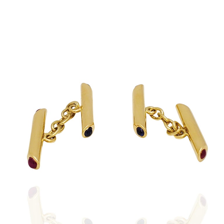 A charming pair of bar cufflinks crafted in 18 karat yellow gold.  The end of one bar is set with cabochon rubies, the other is set with sapphires.  The bars are connected by an oval link chain.  Stamped D G with UK assay marks. Cufflinks are not