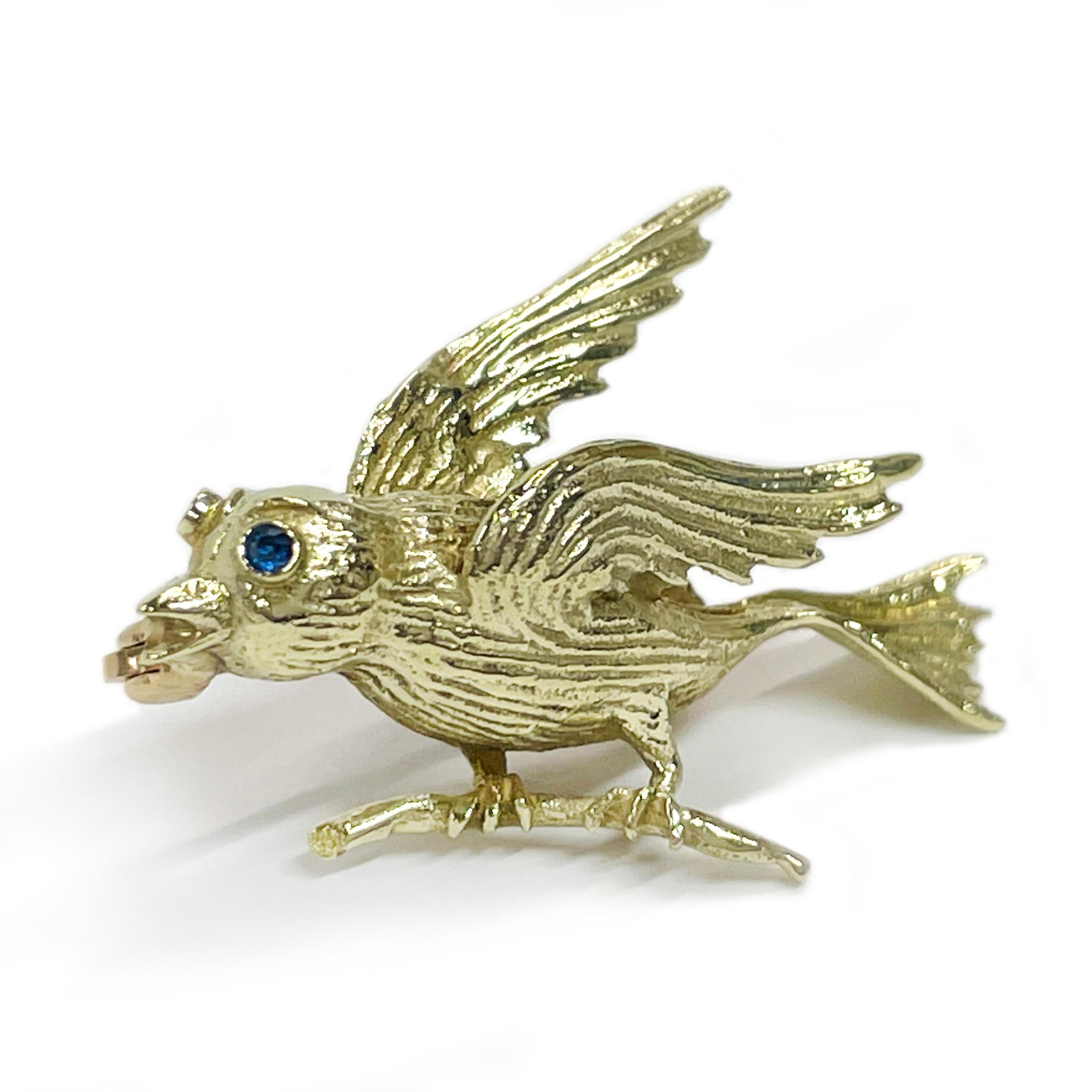 14 Karat Yellow Gold Sapphire Bird Brooch. This adorable little bird is standing on a branch and seems to be ready to take flight. The body features a textured body, wings, feet and head with a round blue sapphire flush-set as the eye. The blue