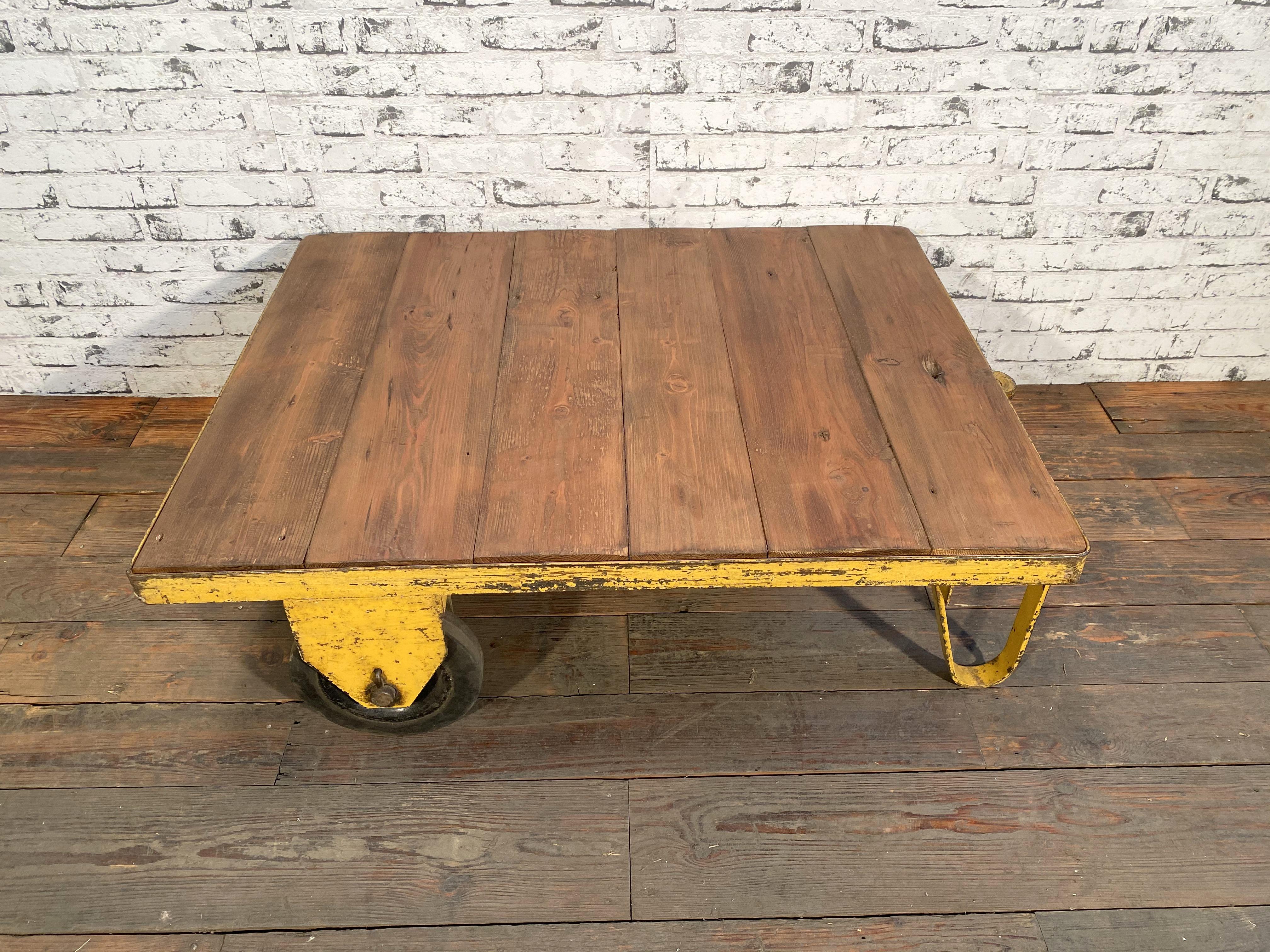 Former pallet truck from a factory now serves as a coffee table. It features a yellow iron construction with two wheels and a solid wooden plate. The weight of the table is 30 kg.