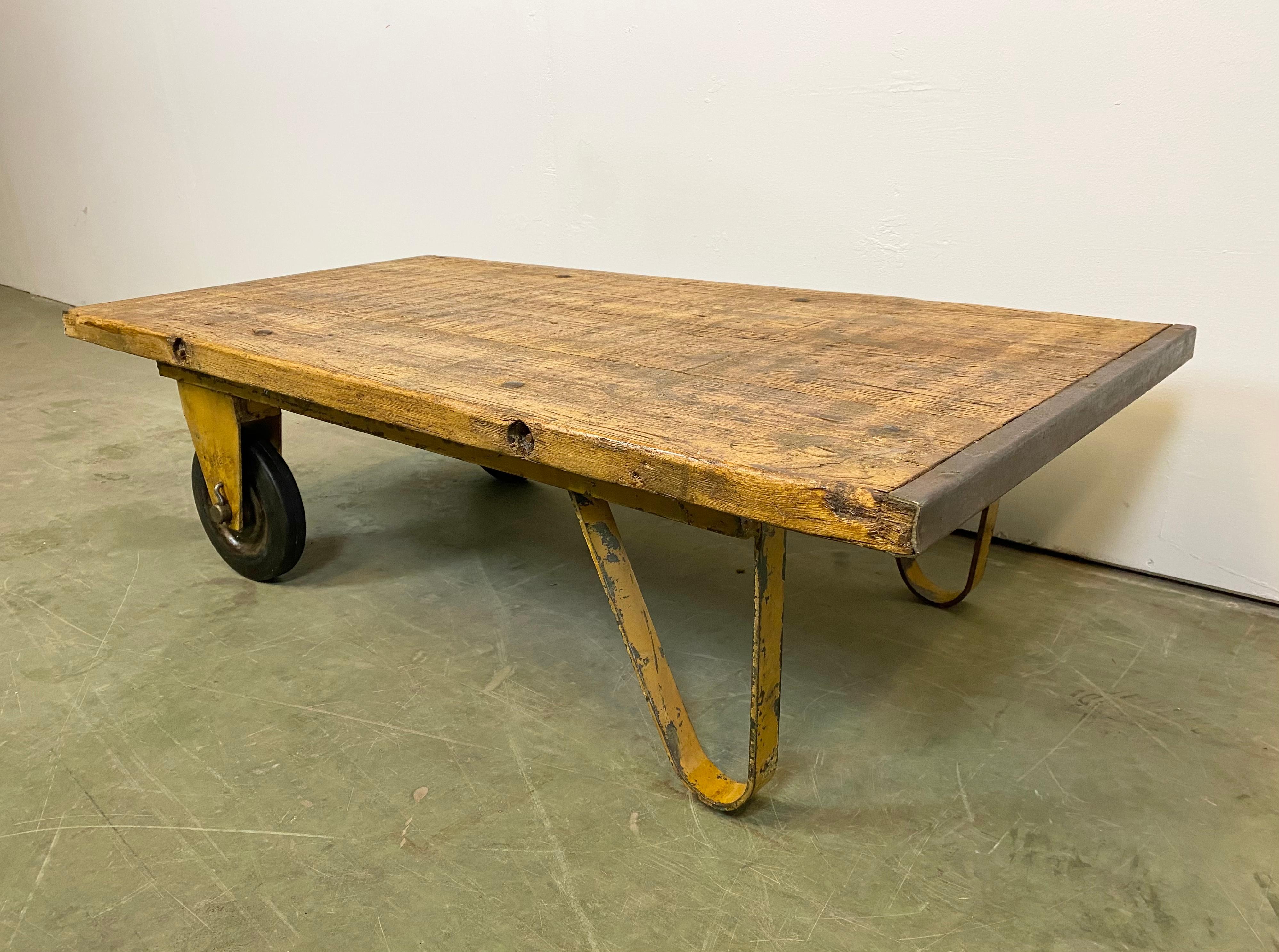 Former pallet truck from a factory now serves as a coffee table. It features a yellow iron construction with two wheels and a solid wooden plate. The weight of the table is 33 kg.