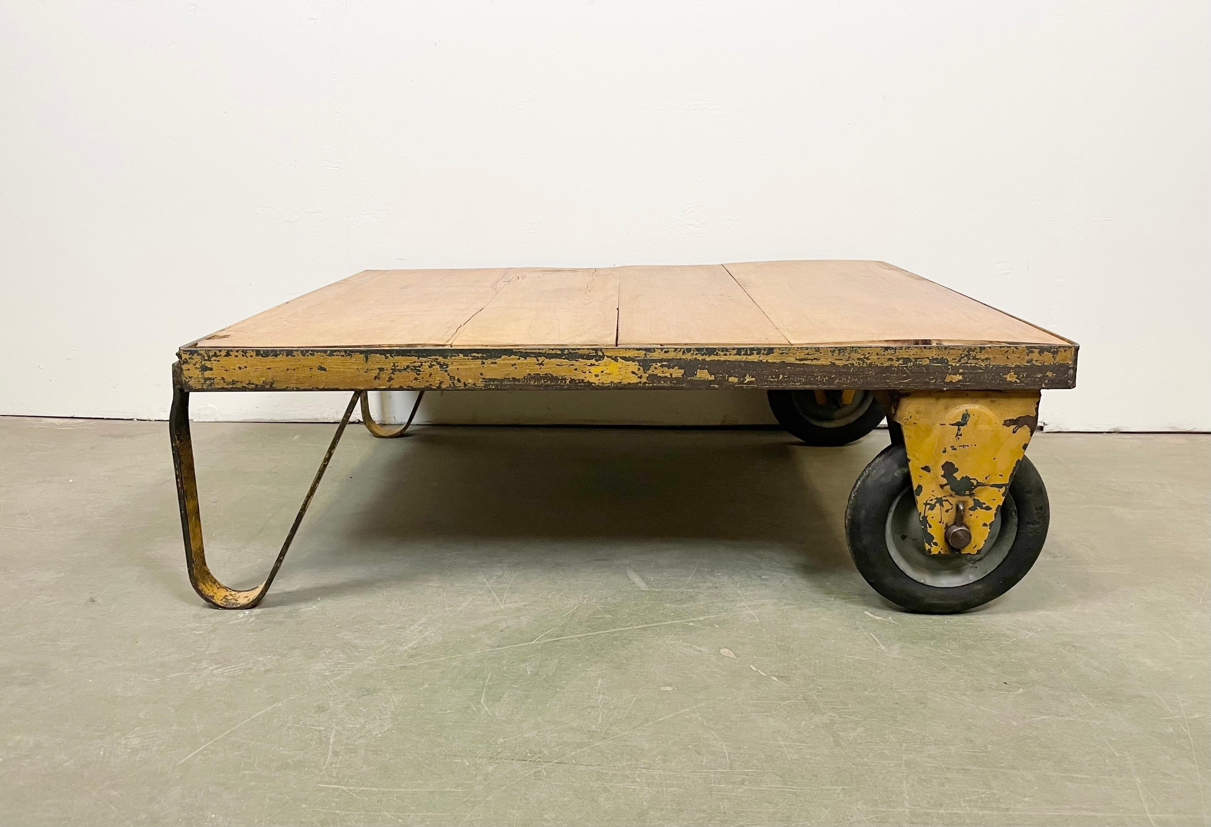 Former pallet truck from a factory now serves as a coffee table. It features a yellow iron construction with two wheels and a solid wooden plate. The weight of the table is 35 kg.