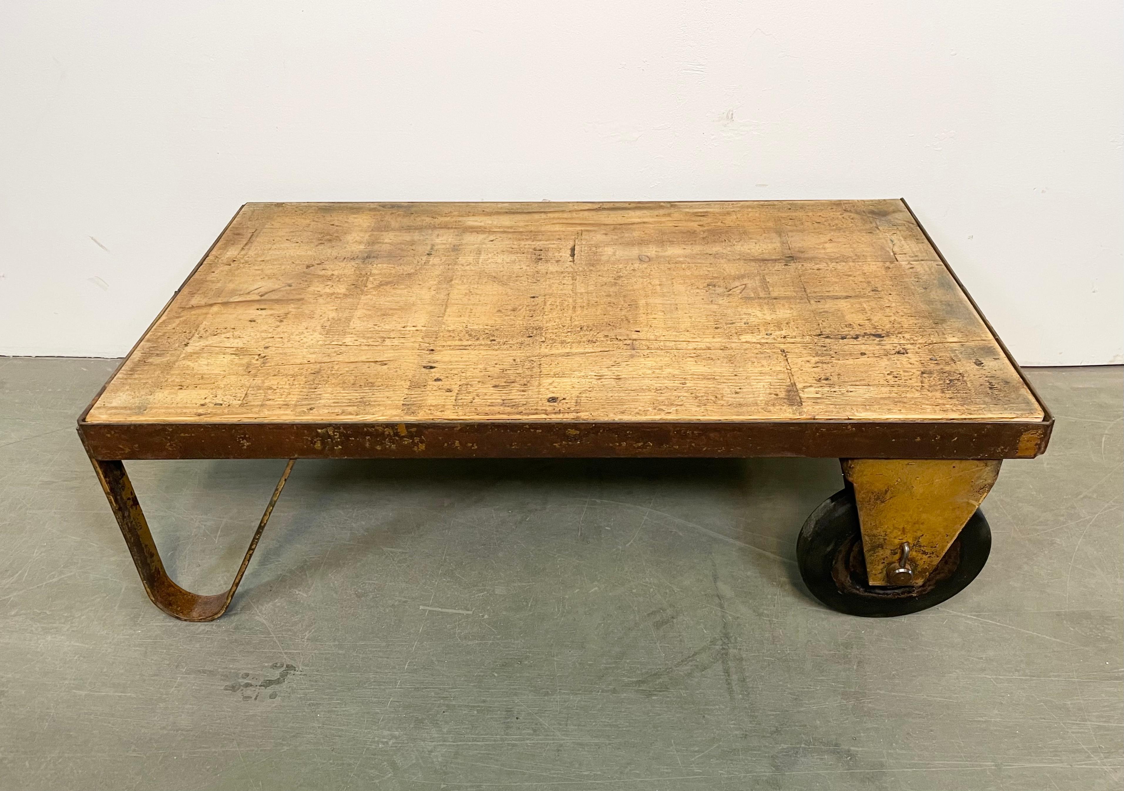 Former pallet truck from a factory now serves as a coffee table. It features a yellow iron construction with two original wheels and a solid wooden plate. The weight of the table is 25 kg.