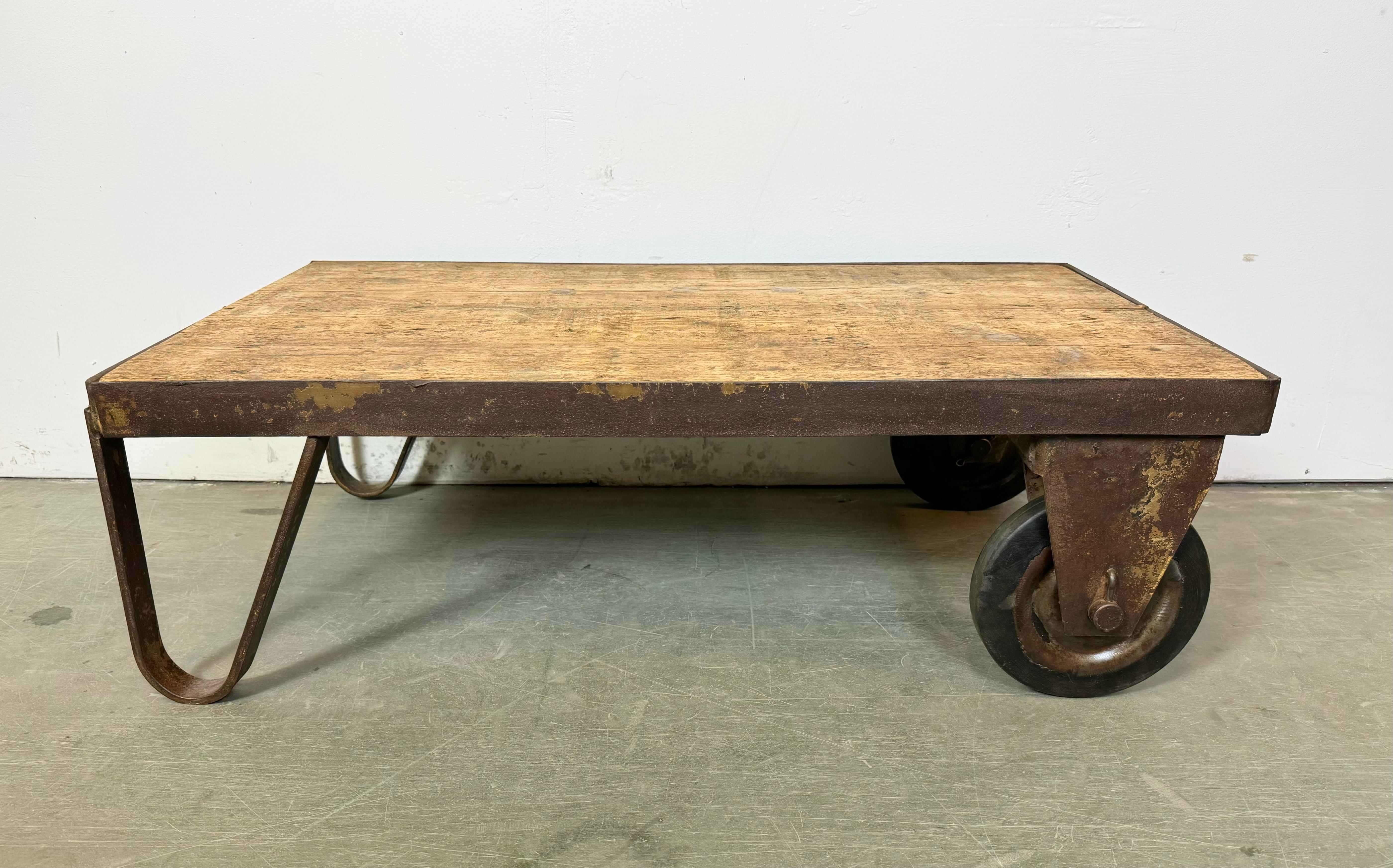 Former pallet truck from a factory now serves as a coffee table. It features a yellow iron construction with two original wheels and a solid wooden plate. The weight of the table is 25 kg.