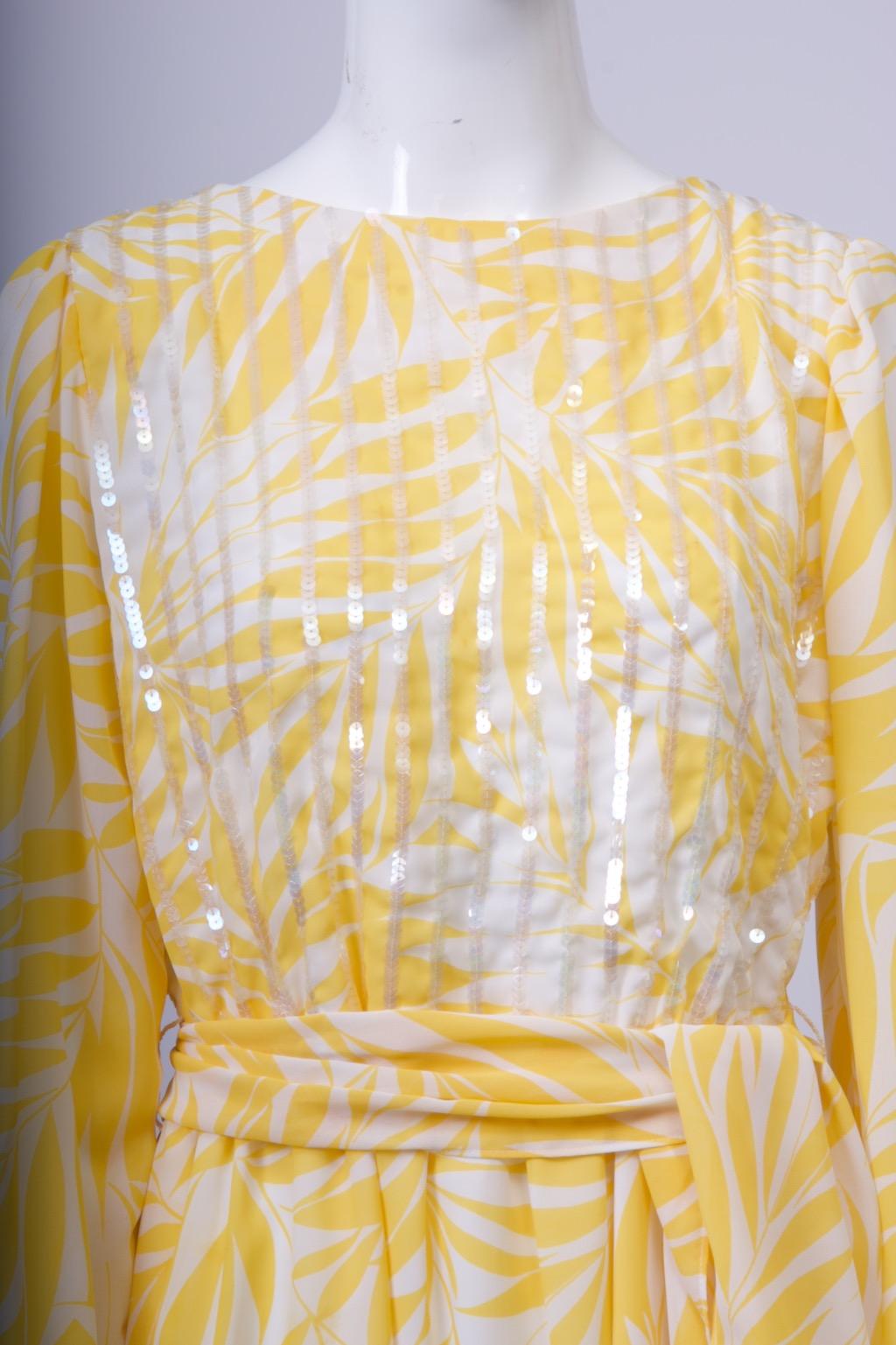 A summery 1970s cocktail dress in polyester chiffon print of yellow leaf design on a white ground, the round-neck bodice embellished with vertical stripes of iridescent sequins. 
There is slight gathering at the waistline, below which the semi-full