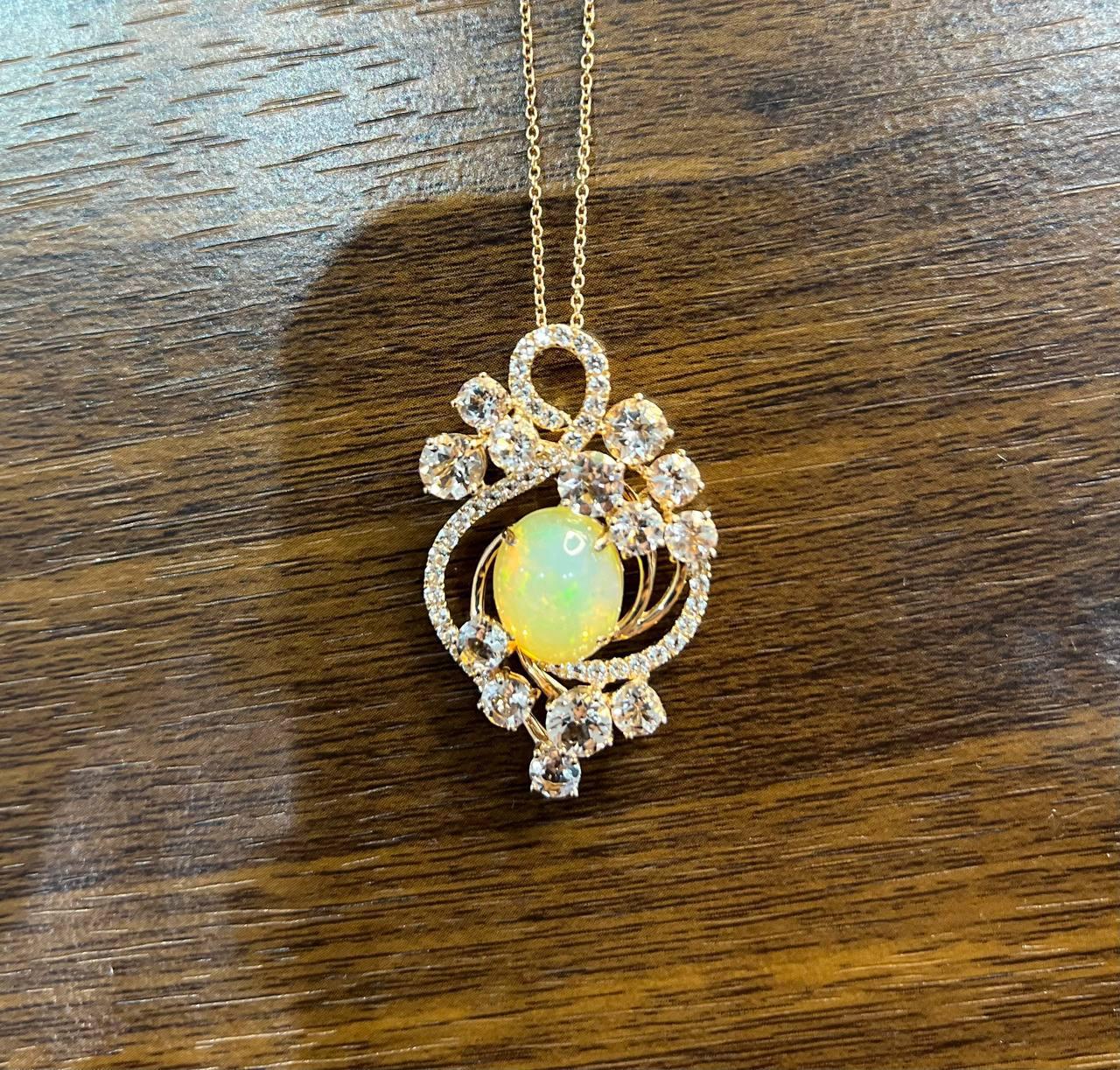This elegant pendant features a central opal cabochon surrounded by a swirl of sparkling round-cut diamonds and peach morganites. The intricate design includes floral motifs, enhancing the opal's vibrant play of color. The pendant is set in a
