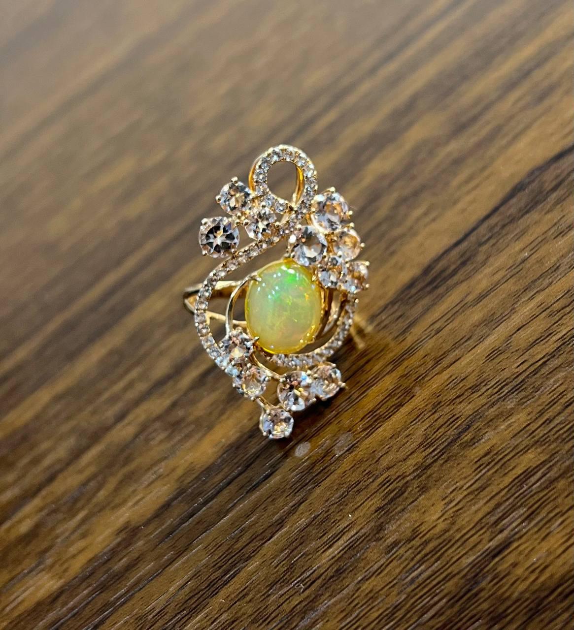 Elegante anillo de ópalo, morganitas y diamantes. Elaborada en oro rosa de 14 quilates, esta exquisita pieza presenta un ópalo central con un cautivador juego de colores. Alrededor del ópalo hay brillantes diamantes dispuestos en un intrincado