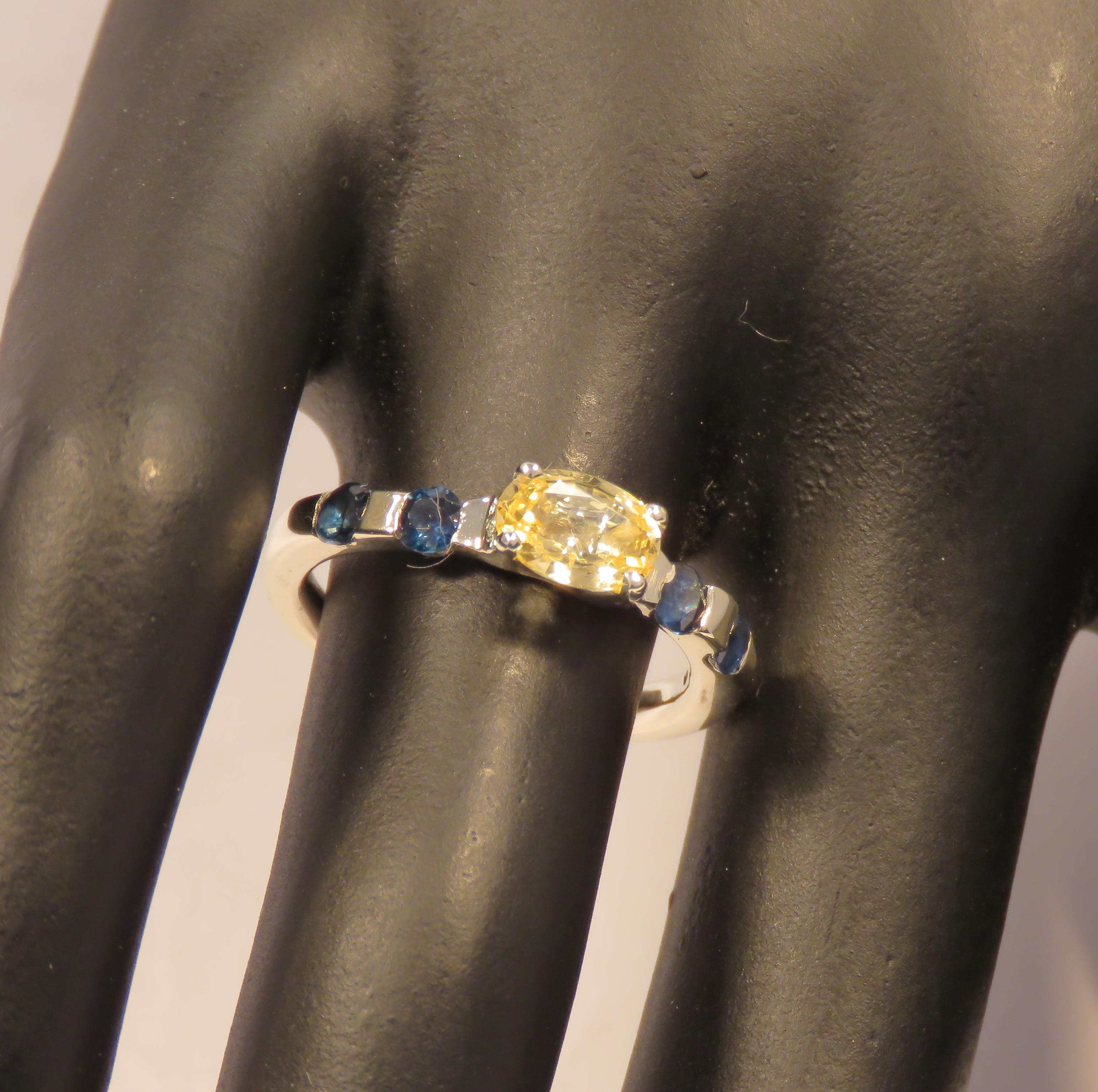 Modern gracious ring with a centre oval yellow sapphire weighing 1.00 ctw, and two natural brilliant cut blue sapphires weighing 0.30 ctw either side of the setting. The ring is completely handcrafted in 9 karat white gold. The dimension of the