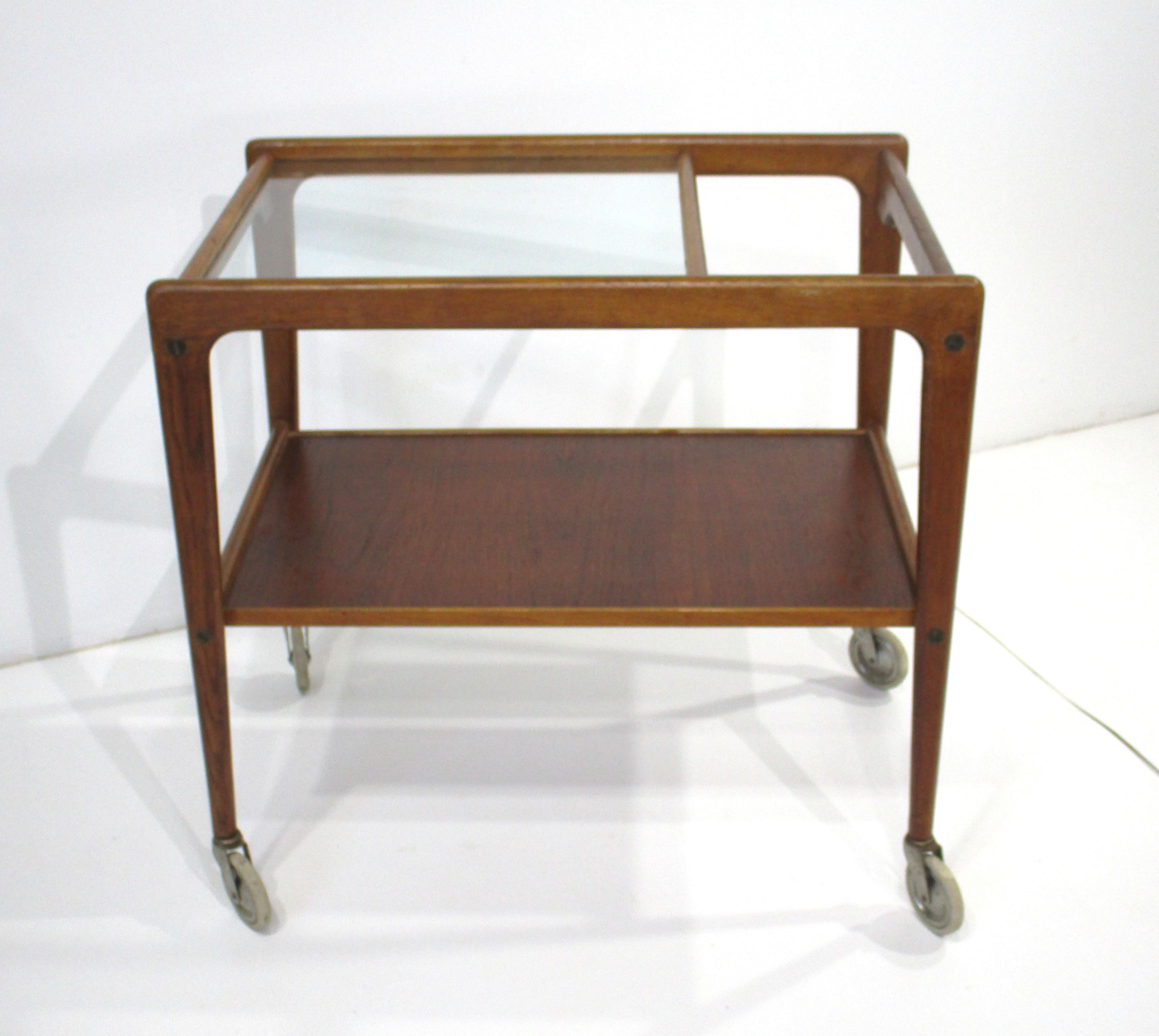 Chariot de bar roulant fonctionnel en bois de teck avec plateau partiel en verre, l'autre côté présentant un espace qui forme la zone de la poignée. Ce chariot fin et effilé, doté de petites roues et d'une esthétique simple, a été conçu par l'un des