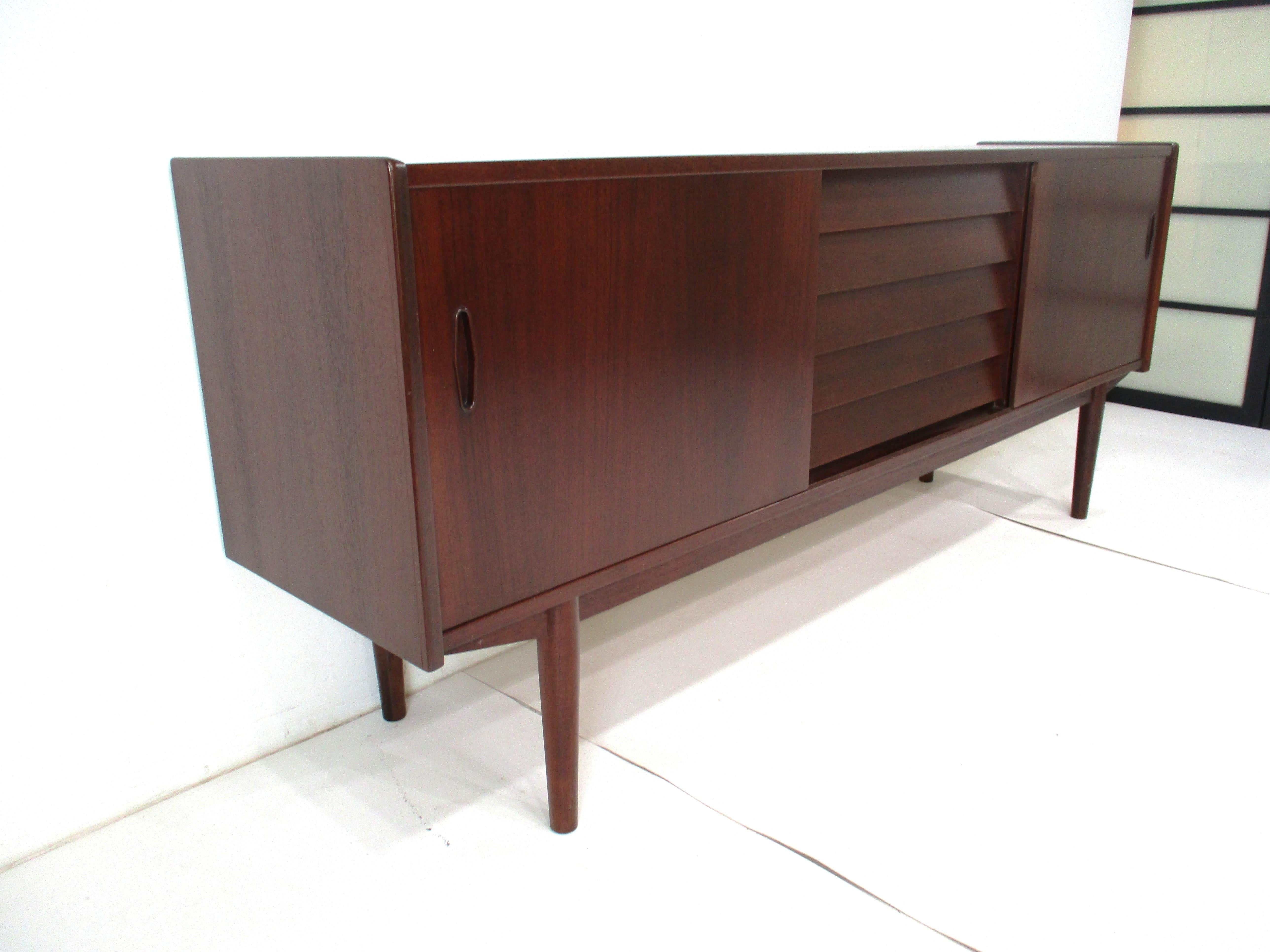 Una credenza / servidor de madera de teca oscura muy bien trabajada con cinco cajones centrales el superior con ranuras divididas y fondo de fieltro . A cada lado, una puerta corredera con estantes ajustables de madera de abedul clara en contraste,