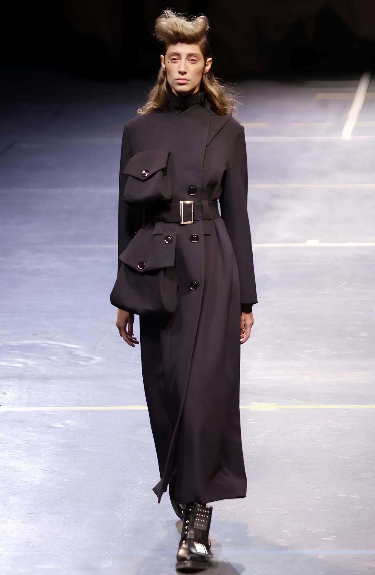 Ein strukturiertes schwarzes Wollkleid mit asymmetrischer, gewickelter Vorderseite und einem hohen Kragen mit Schnalle. Der doppelreihige Verschluss ist dezentral angeordnet und wird von übergroßen, glänzenden Knöpfen unterstrichen. Ein breiter