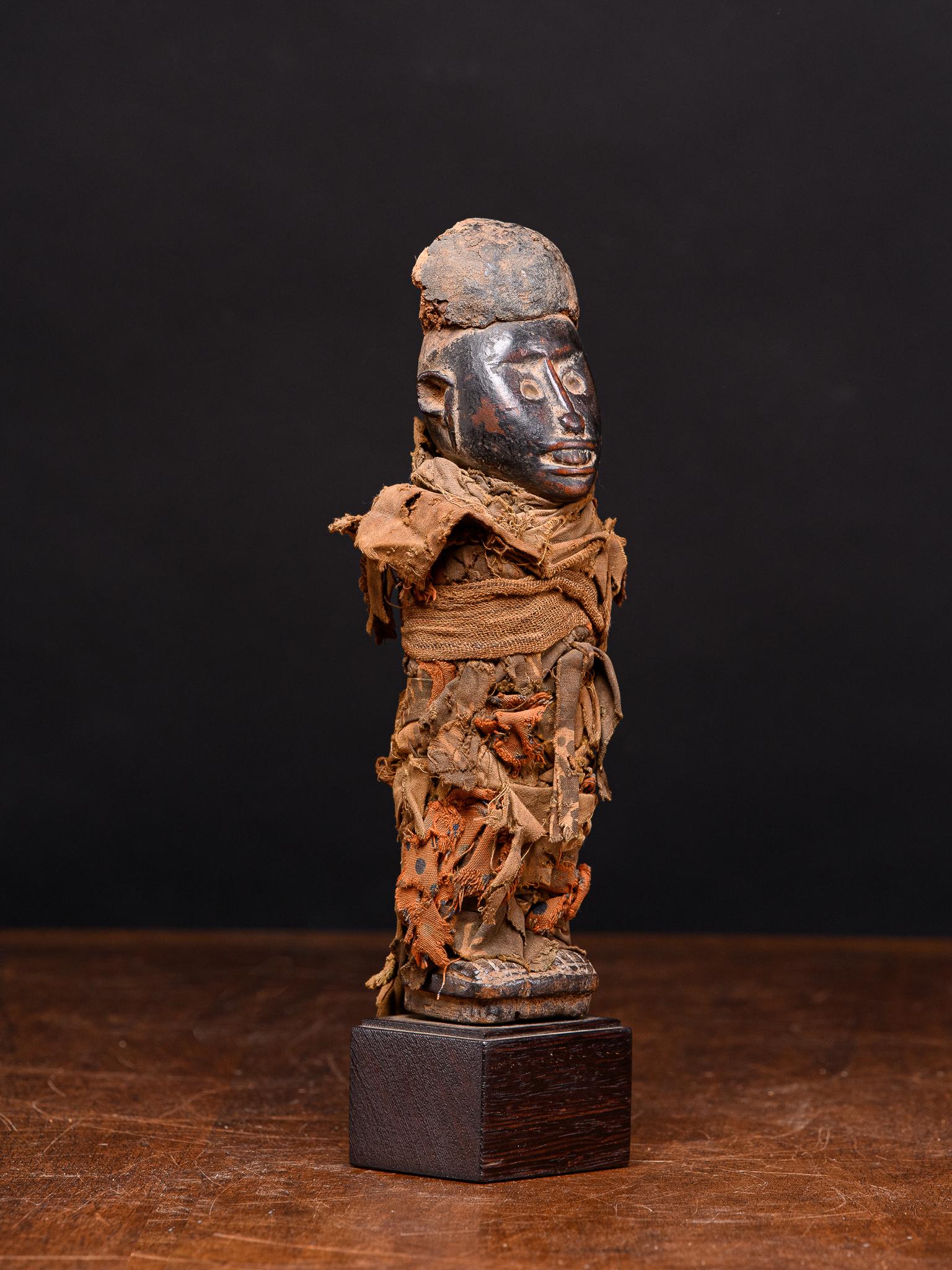 The statue is standing on a rectangular base, the body covered with bundles of knotted cloth from feet to chin, the rounded, ovoid head with tight mouth baring teeth and a slender nose bisecting the circular eyes inlaid with glass and wearing a tall