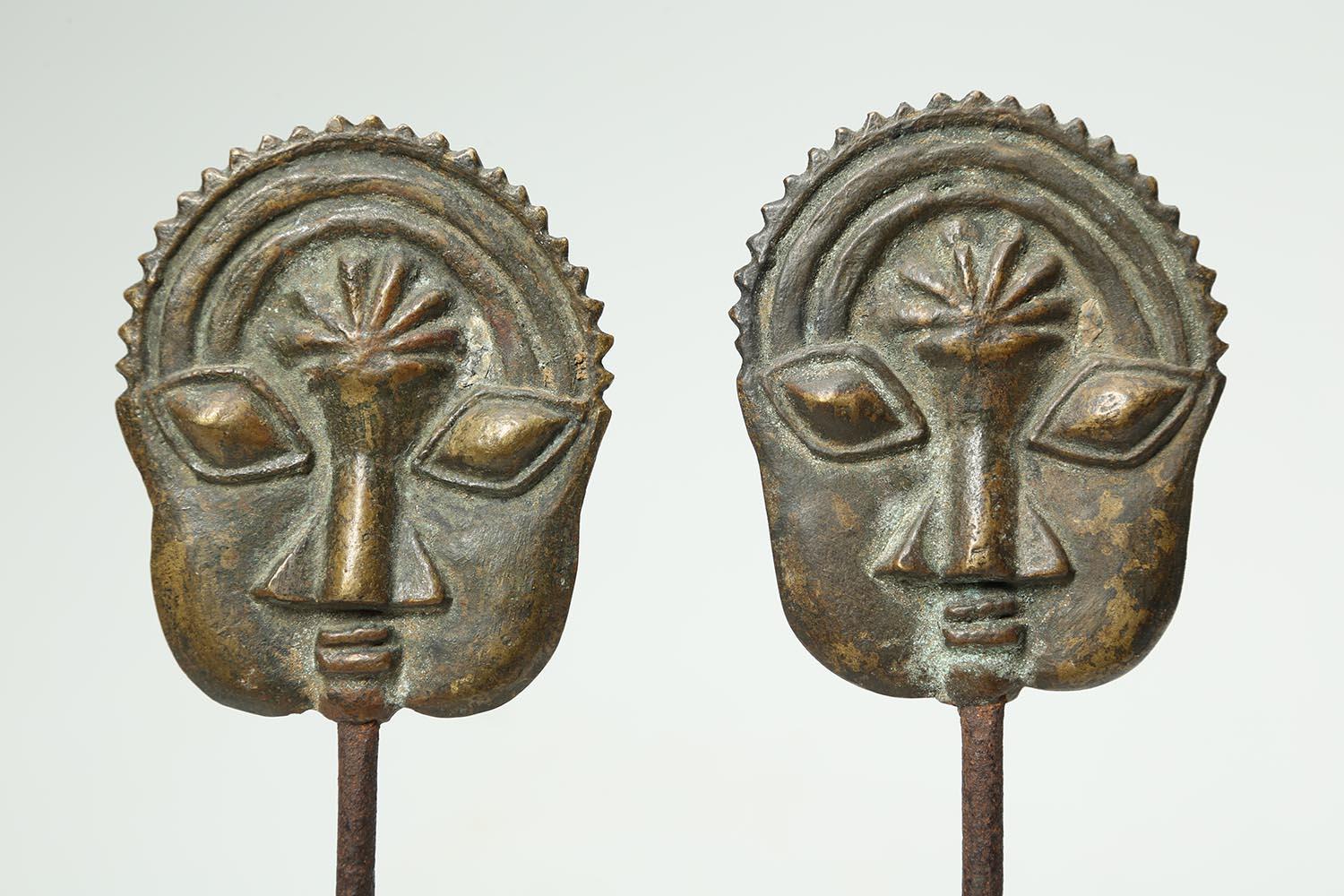 Yoruba People, Ogboni men's secret society pair of brass pins with expressive faces, Nigeria, early 20th century. A pair of cast brass or bronze Ogboni society pins with matched flat faces on iron pins, mounted on custom base. Each of the faces 3