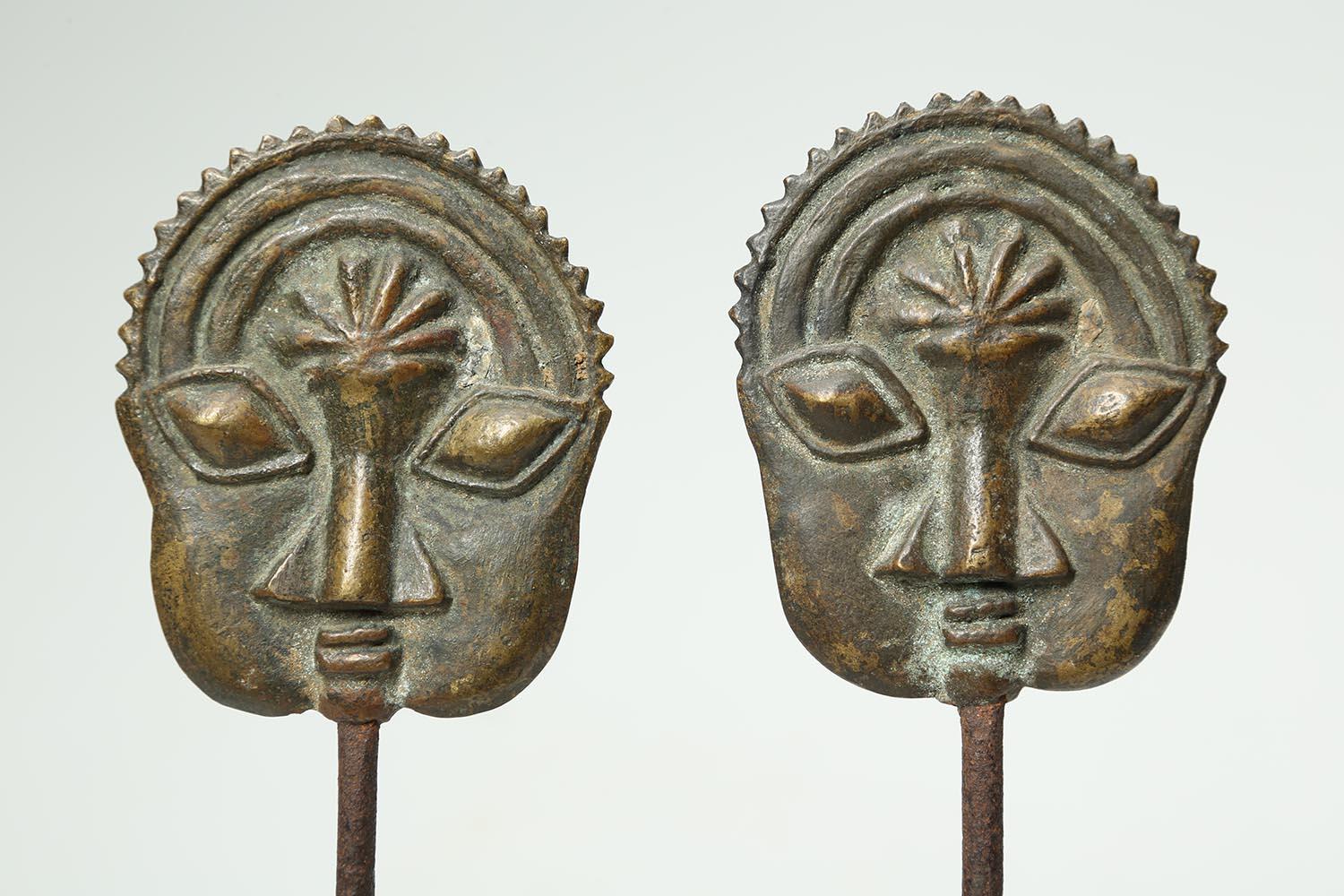 Yoruba people, Ogboni men's secret society pair of brass pins with expressive faces, Nigeria, early 20th century. A pair of cast brass or bronze Ogboni society pins with matched flat faces on iron pins, mounted on custom base. Each of the faces 3