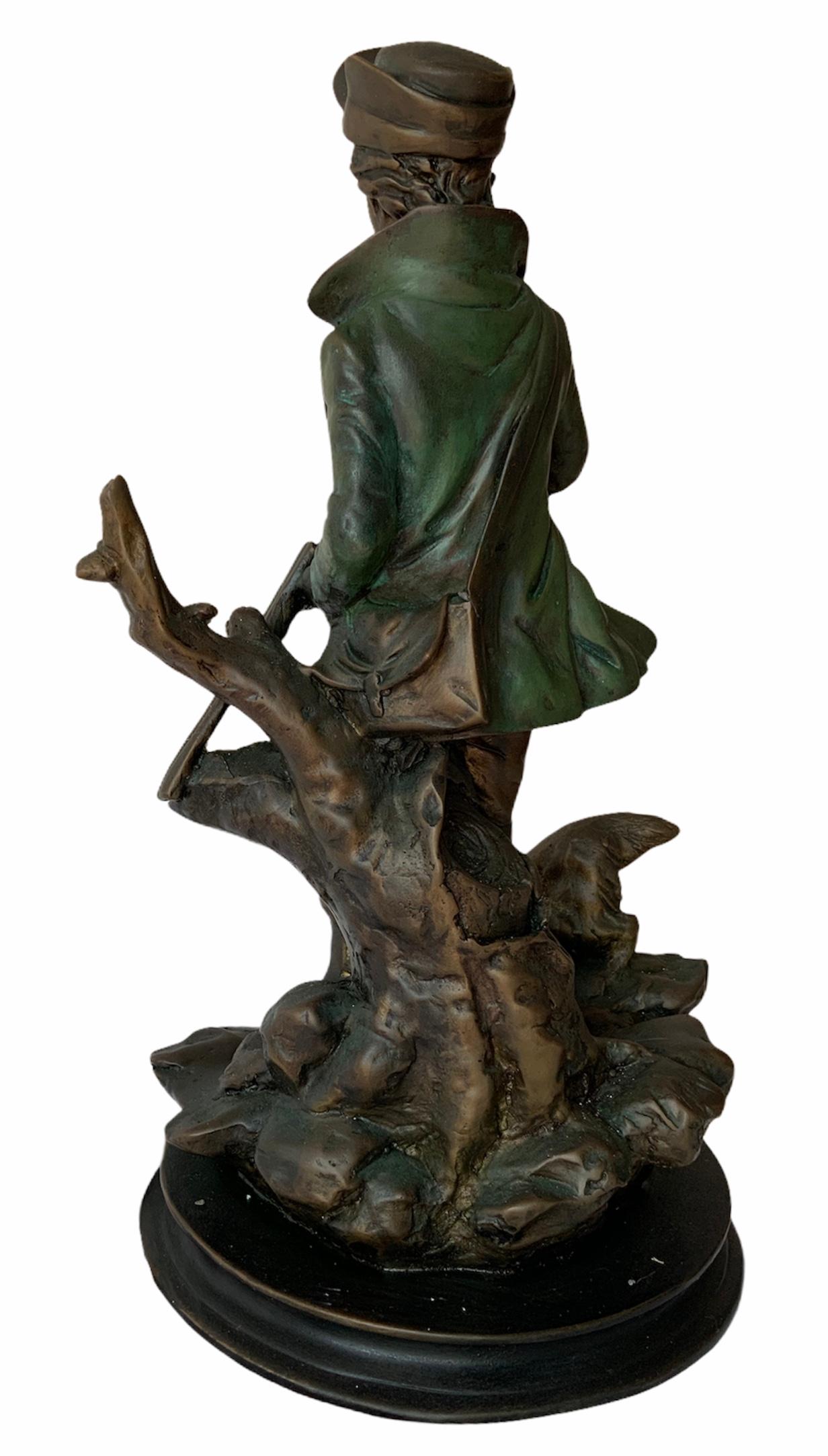 This a metal sculpture of a young man hunter with his dog. He is dressed with an open green coat, long pants, pair of boots, a hat and a bullet belt around his waist. It appears that he is loading his rifle. He is standing in front of a cut tree