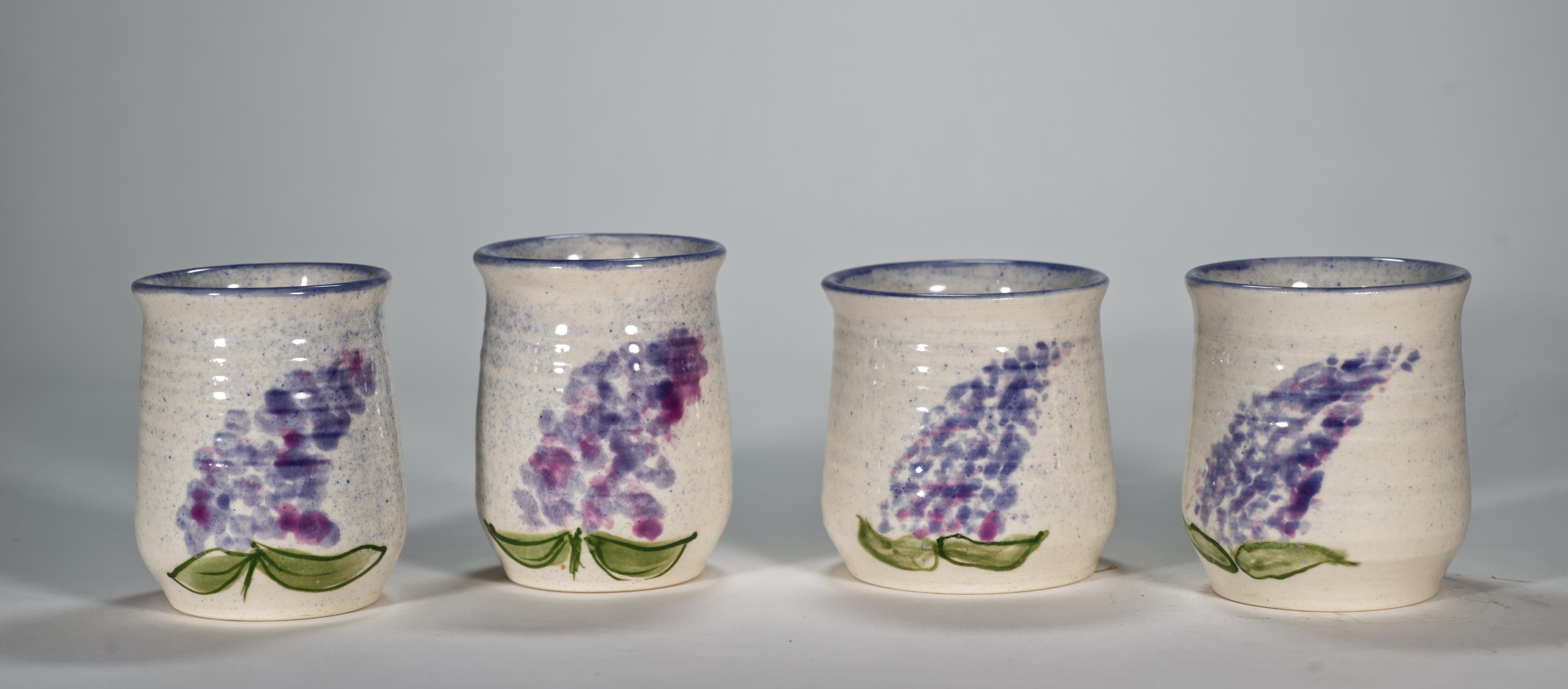  Charmant ensemble vintage de 4 verres en céramique présentant un motif peint à la main d'une branche de lilas en fleurs dans des couleurs pastel sur un fond émaillé blanc cassé et bleu. Les bords sont décorés d'une bande bleu clair. Les verres sont