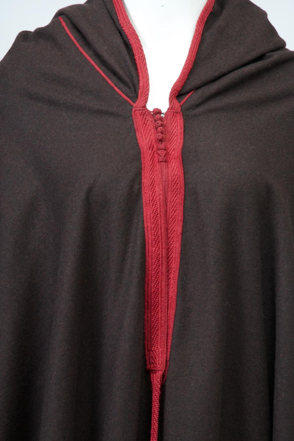 Yves Saint Laurent hard-to-find vintage 1979s cape from the designer’s Moroccan collection. Crafted of black wool, the simply cut long cape features a pointed hood and burgundy braided trim on all edges, a single row except for the wider area below