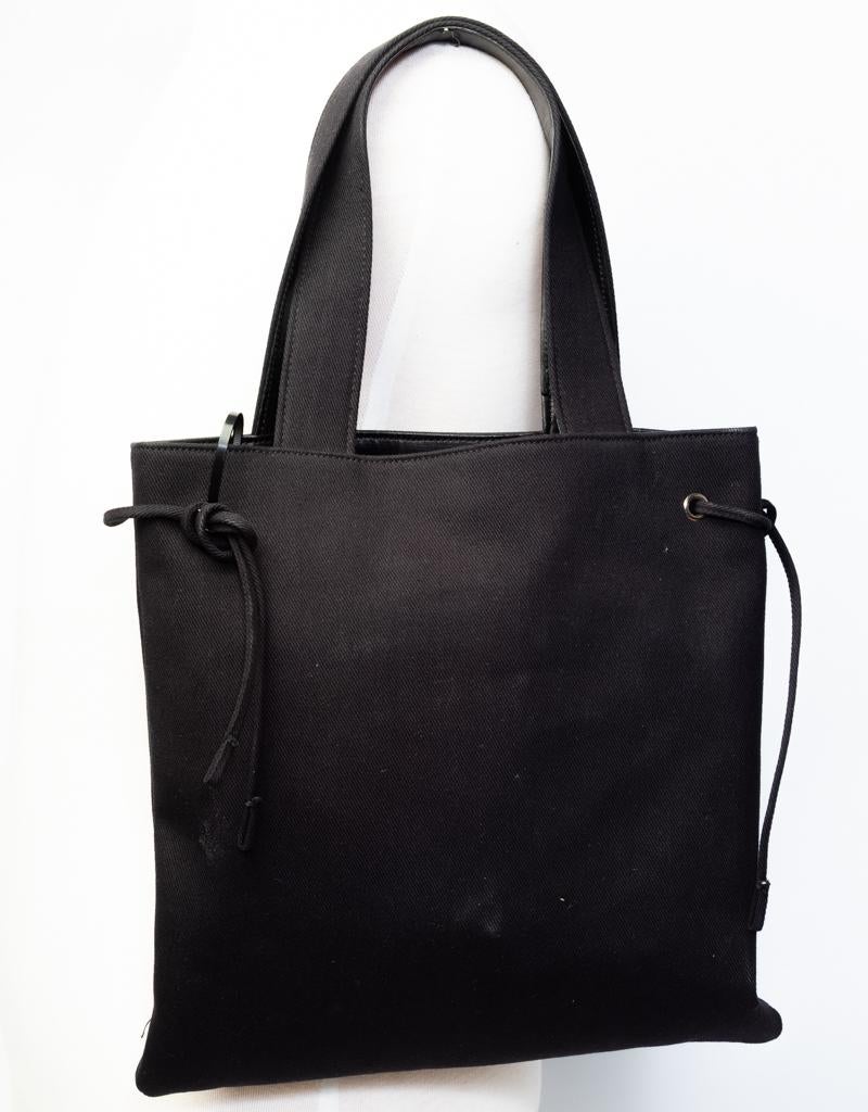 The tote bag features a canvas body with decorative logo sequence embroidery on the front, flat straps with leather details, drawstring and snap closure at top, a satin interior and an interior zip pocket.

COLOR: Black
MATERIAL: Cloth
ITEM CODE:
