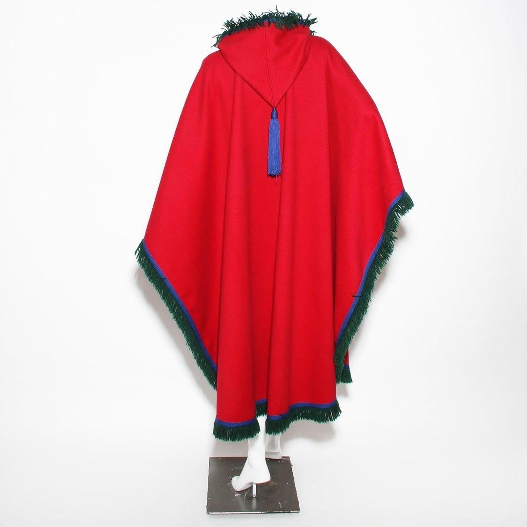 Hooded wool cape by Yves Saint Laurent (Rive Gauche) 
Red wool 
Green fringe 
Blue trim 
Hook and eye neck closure 
Blue rope neck tie 
Hood 
Draped cape no sleeves 
100% wool 
Made in France 
Condition: Excellent condition, little to no visible