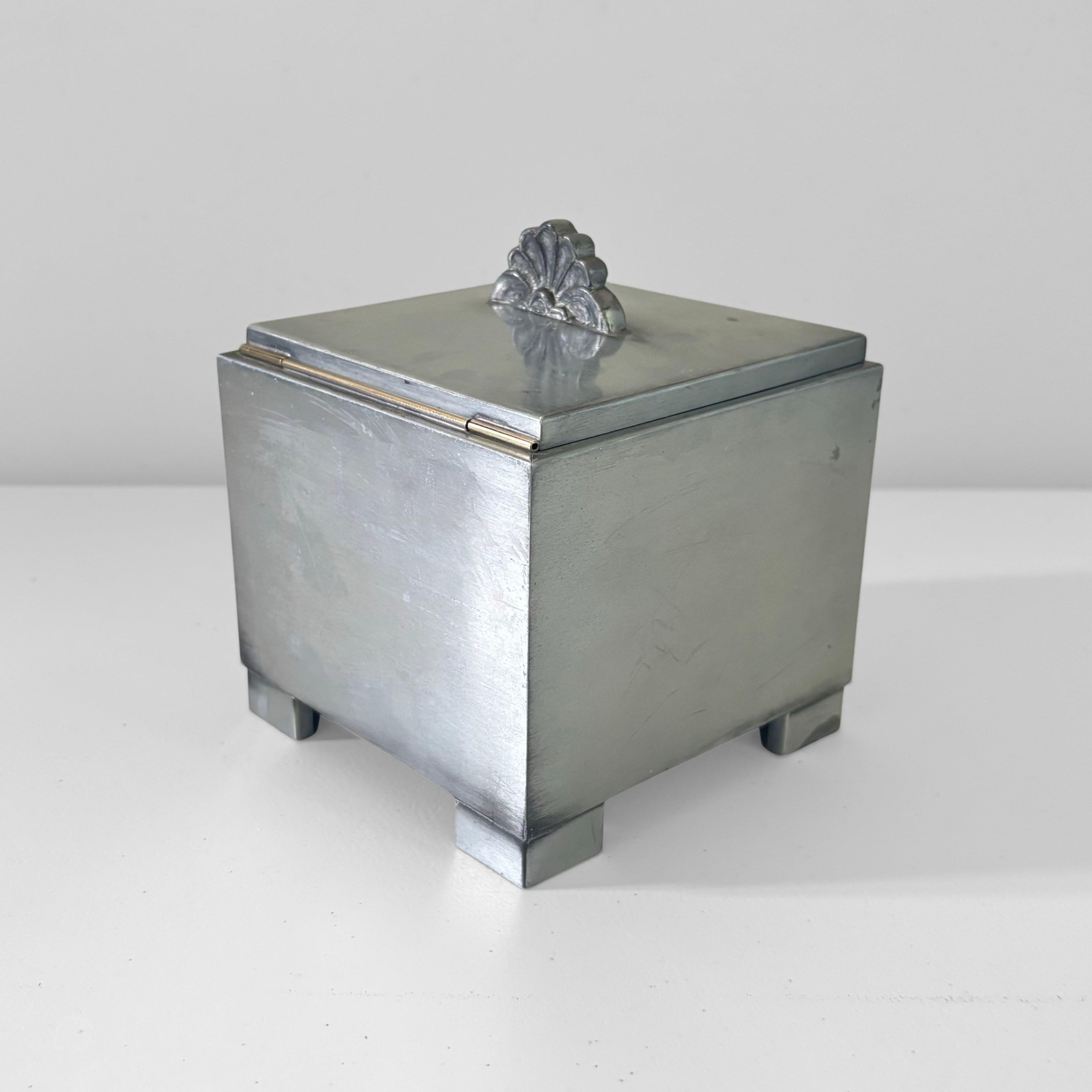 A strict, geometric, cube-shaped Art Deco cigarette box produced by the Swedish manufacturer Ystad Metall in the 1930s. Crafted from pewter, it features a brass hinge for the lid and an interior lined with stained wood. The lid is topped with a