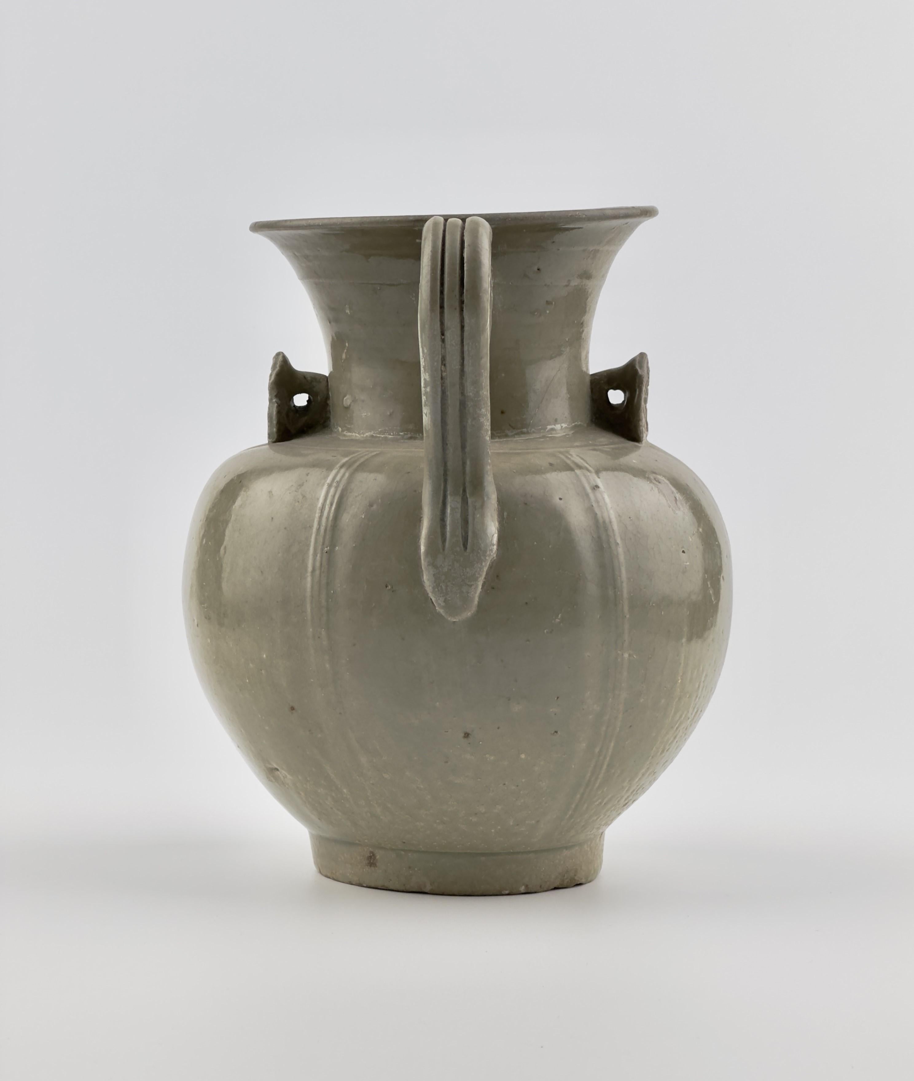 This Yue ware celadon ewer features a gracefully rounded body that expands smoothly, complemented by a wide, flaring mouth. The translucent olive-green glaze envelops the surface, enhancing its depth and highlighting the vertical ridges that run