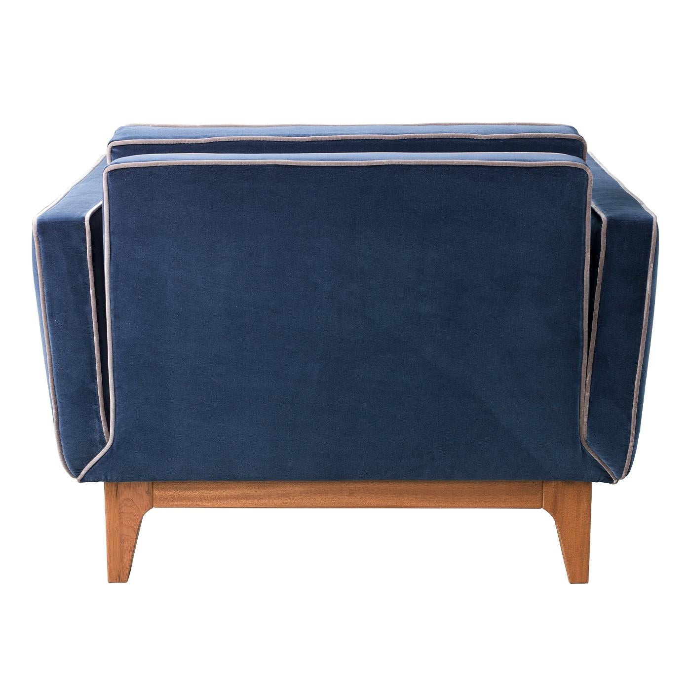 The Yvan Armchair combines mid-century influences with modern aesthetics, boasting a solid mahogany base with a rich brown tone. Its deep blue velvet upholstery is enhanced with contrast trims and classic button-tufted quilting as a subtle nod to