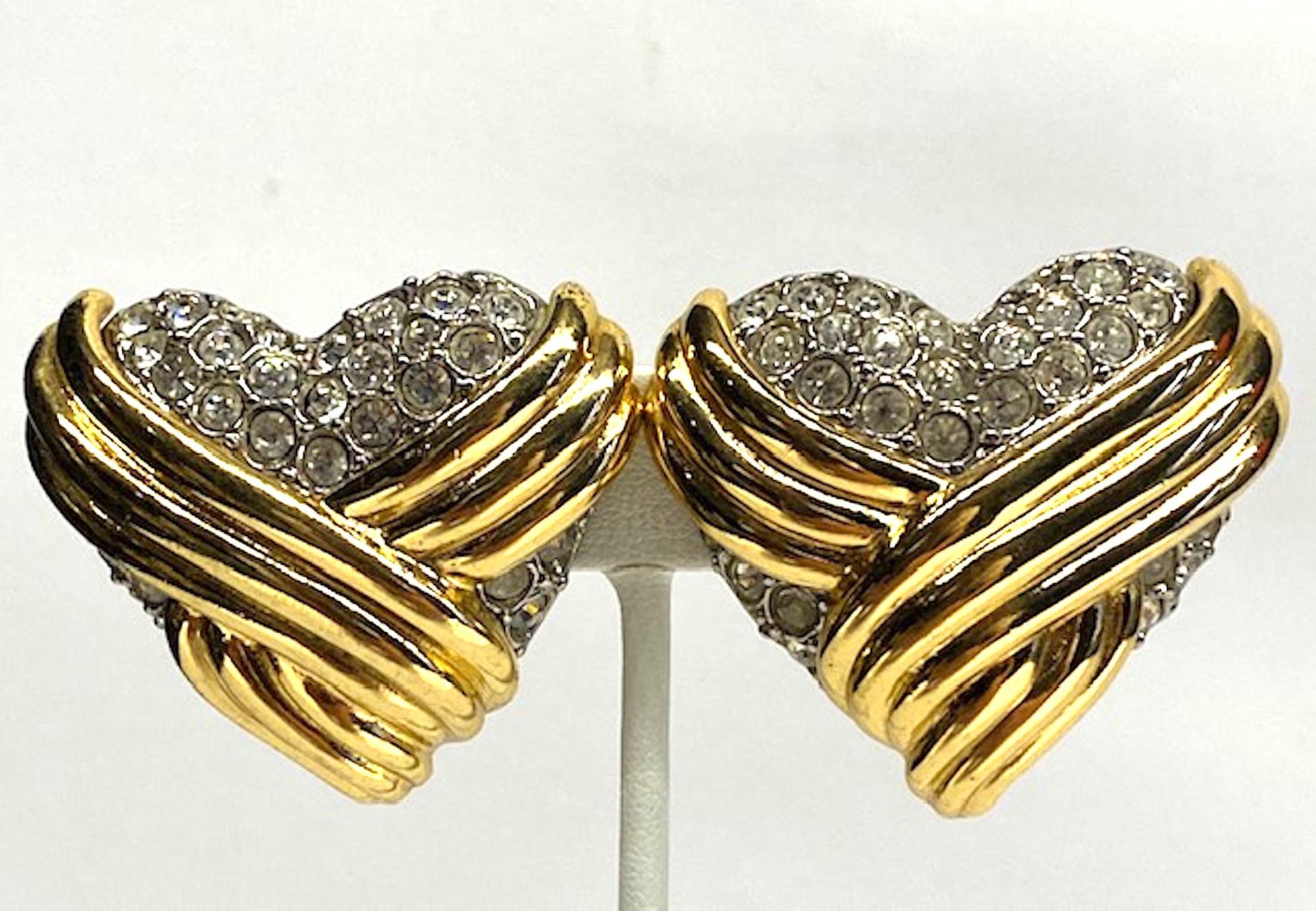A large statement pair of Yves Saint Laurent 1980s heart shape button earrings. The earns are rhodium plated with gold plate ribbed ribbon design, a slight domed shape and round rhinestone accents. Each measures 1.75 inches wide, 1.5 inches high and