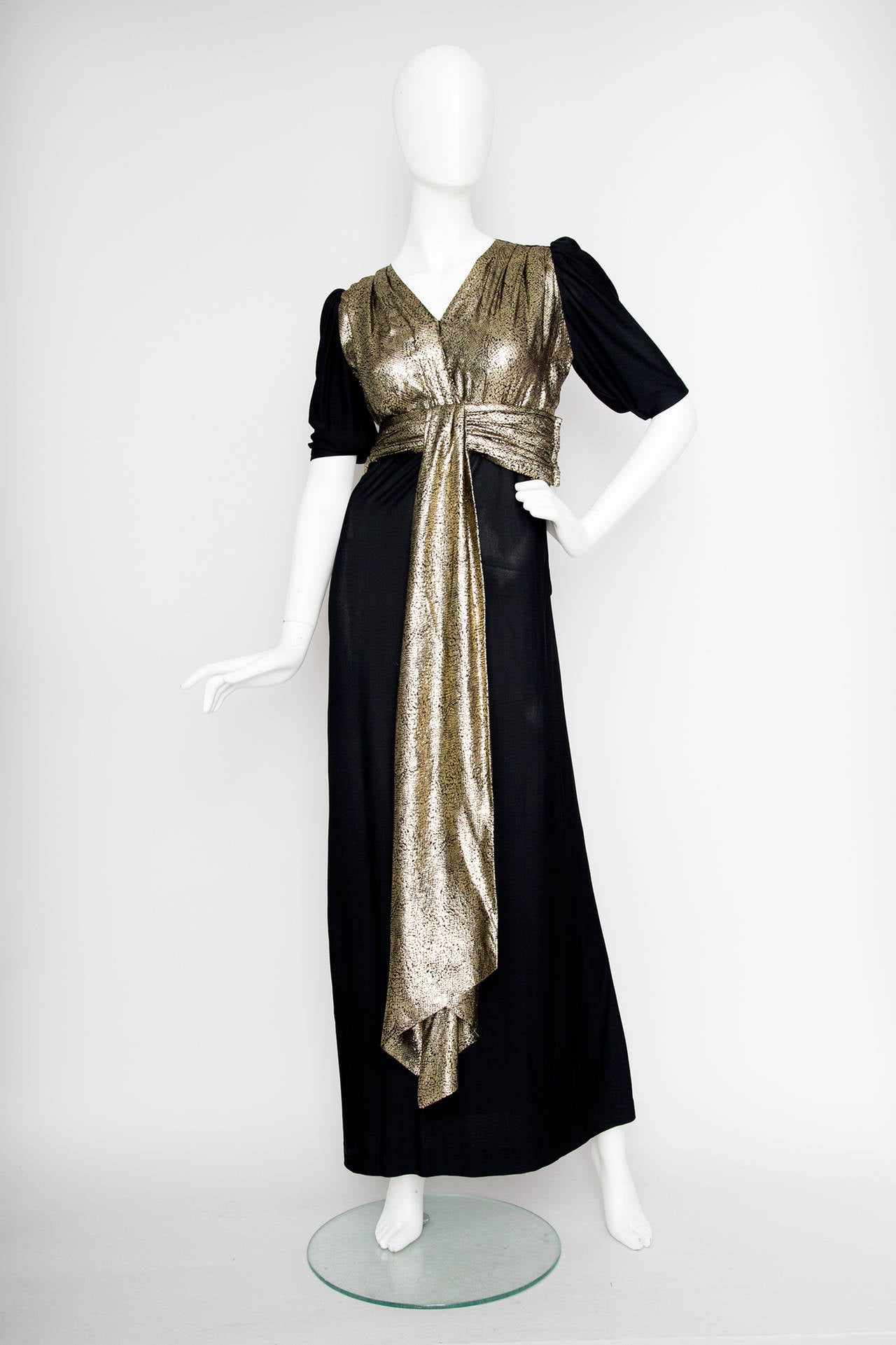A 1970s silk jersey Yves Saint Laurent Rive Gauche black floor length evening dress with a gold-lamé top with a v-neckline, short sleeves and a detachable gold sash gently dropping down in the front of the dress.  

The dress has a zipper closure