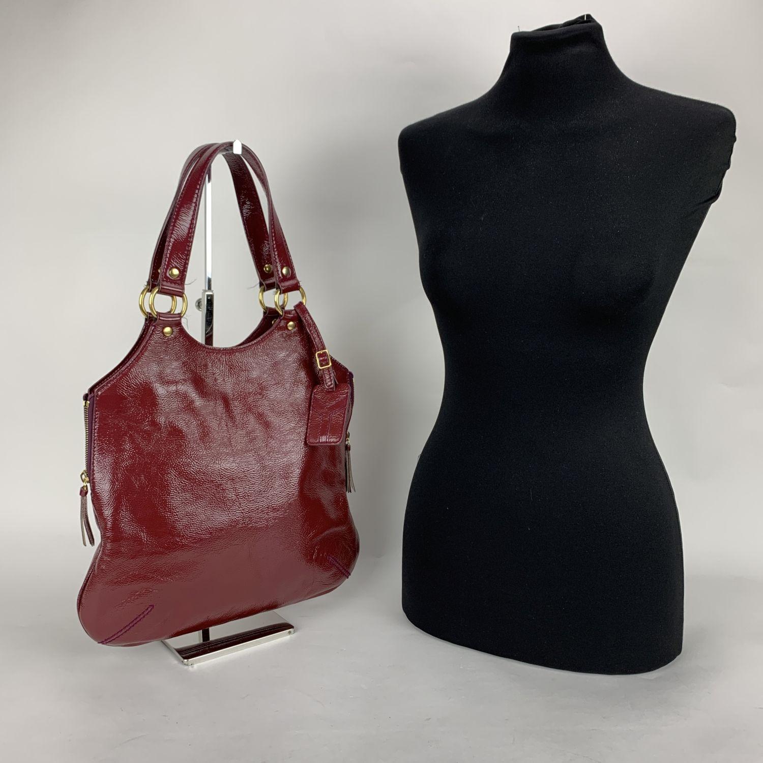 Yves Saint Laurent burgundy 'Metropolis Tribute Bag', crafted in burgundy shiny leather. It features a sleek design in with side zip detailing , an ID tag and gold metal hardware. Magnetic button closure on top. Black satin lining. 1 side zip pocket