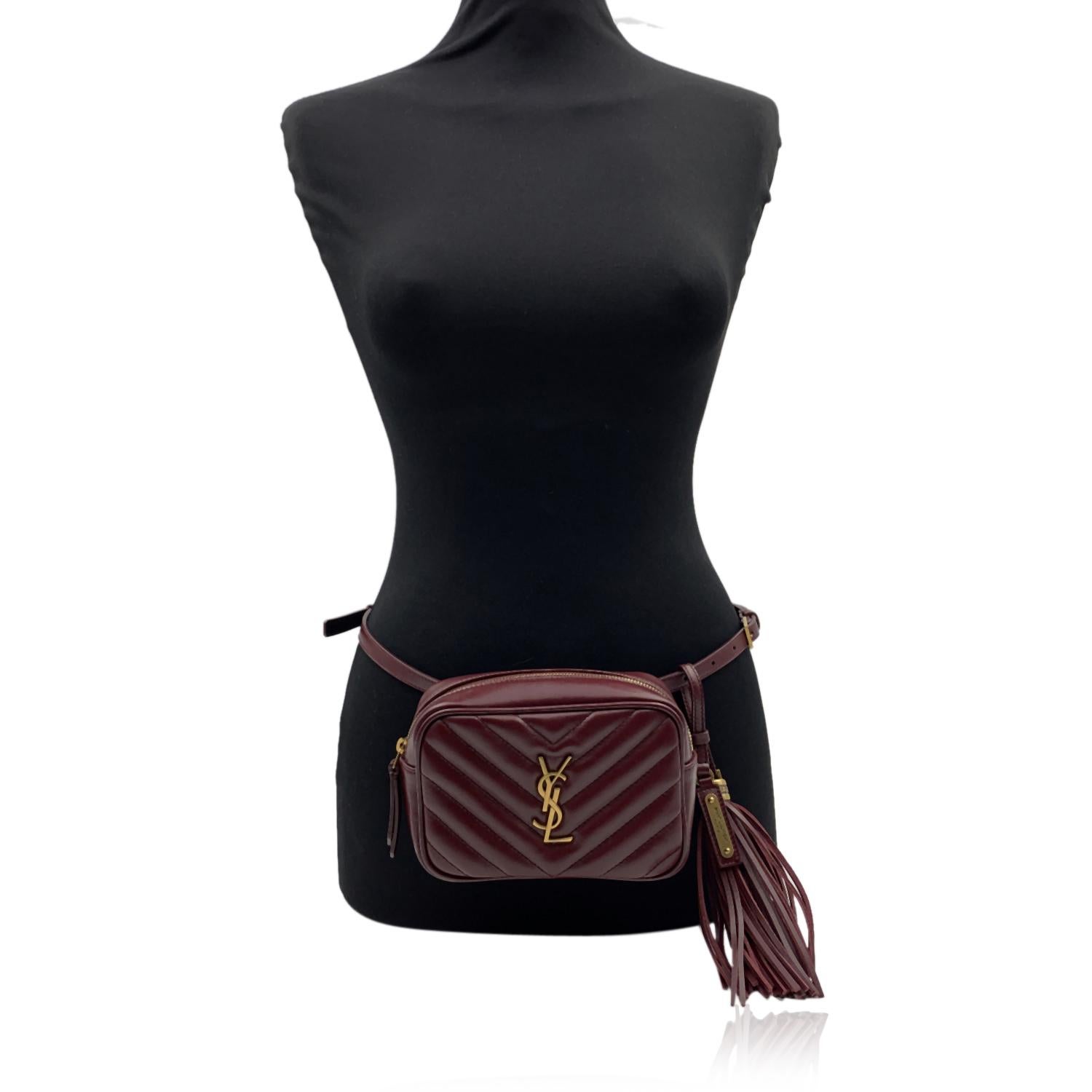 YVES SAINT LAURENT 'Lou' belt bag in burgundy quilted leather. The bag features a v-quilted pattern, gold metal YSL logo on the front and a removable leather tassel. Adjustable burgundy belt (can be adjusted from 65-110 cm). 1 zip pocket on the
